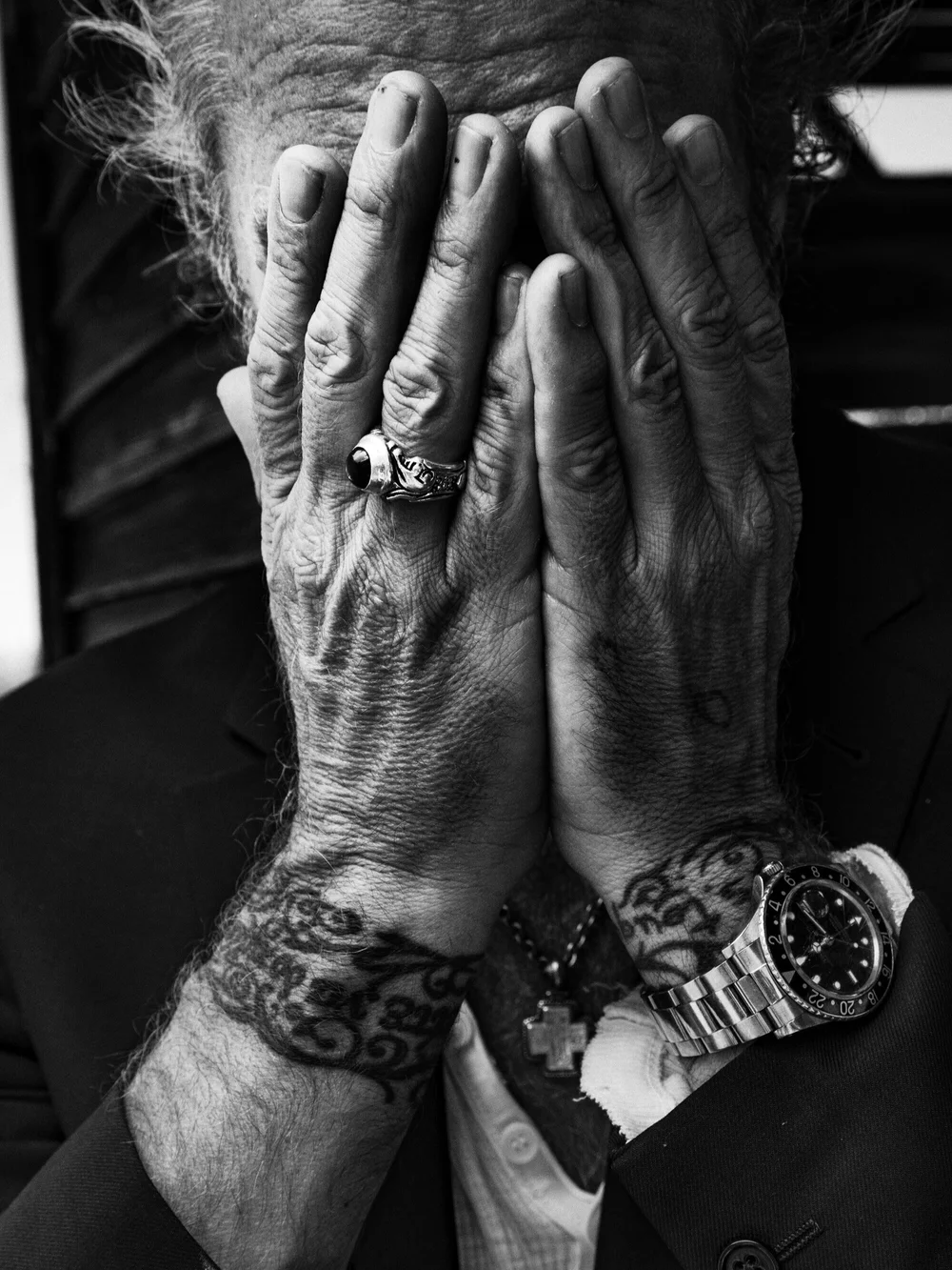 A person with tattoos covering their arms and wearing a watch and a ring, covering their face with their hands.