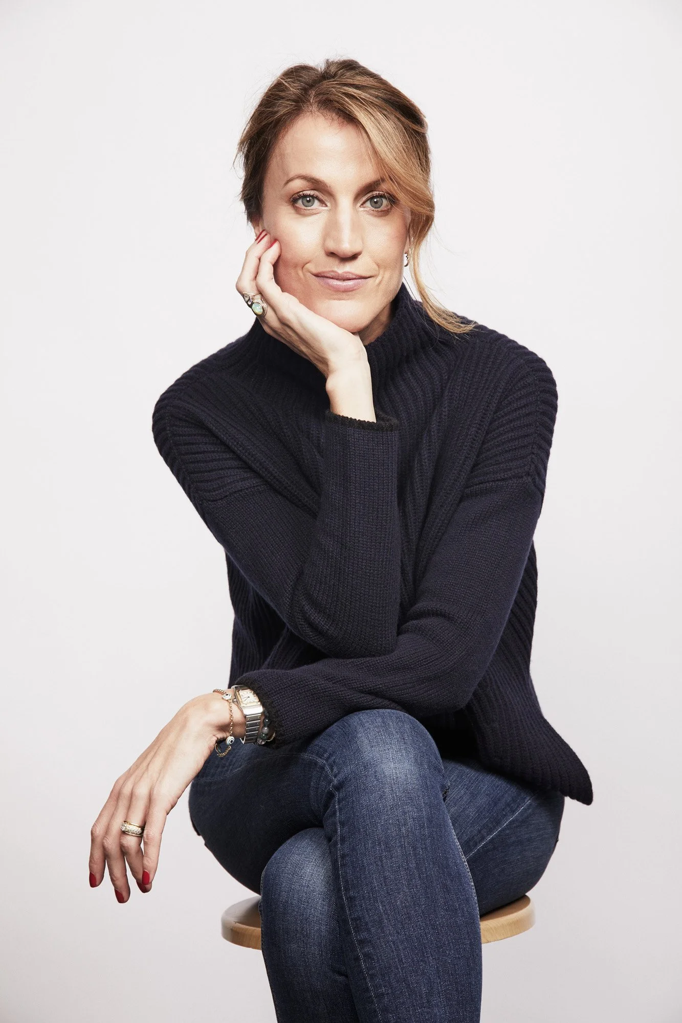 Portrait of a woman with short hair wearing a dark sweater and jeans, sitting on a stool against a plain white background.