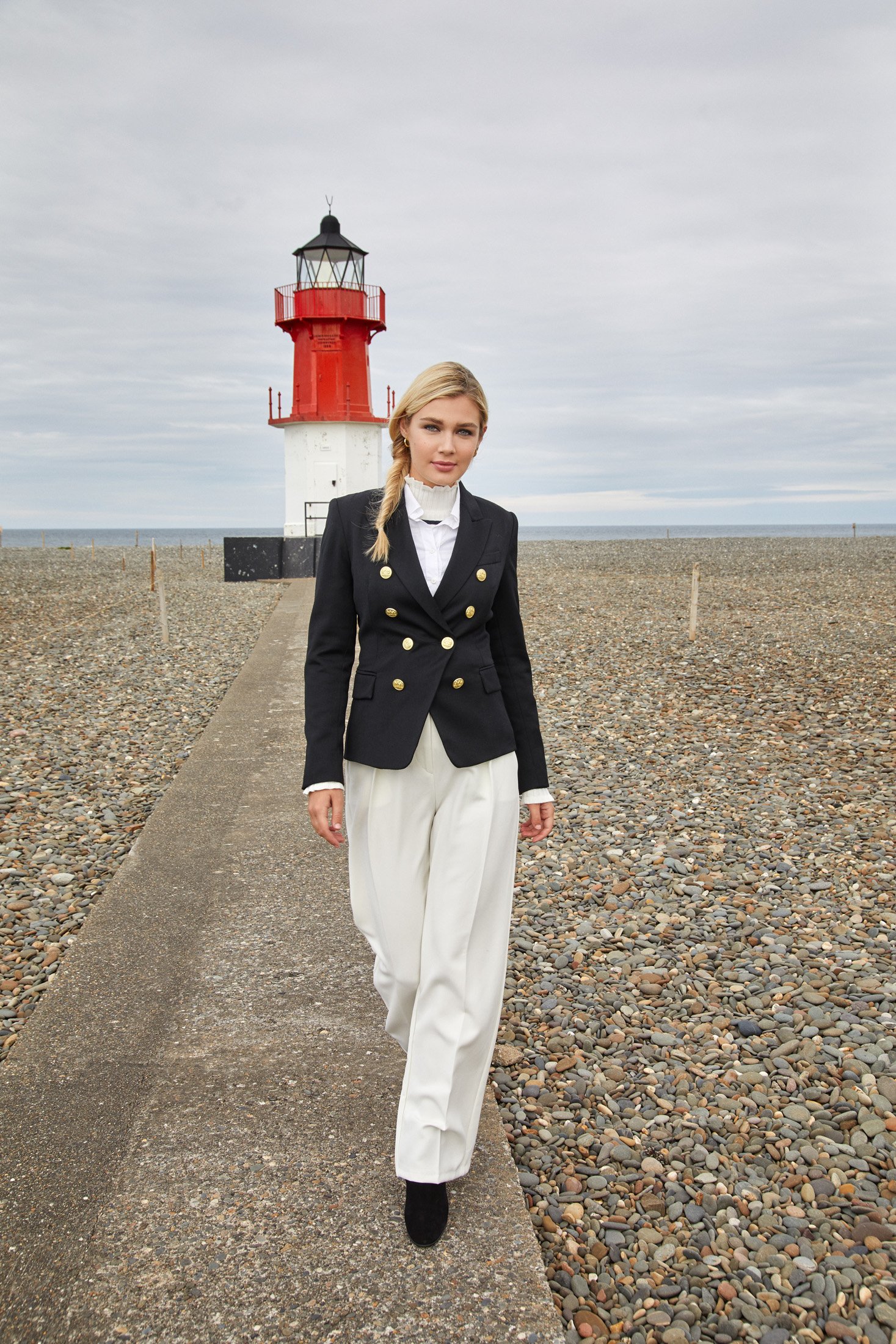 A young woman with blonde hair styled in a side braid, wearing a black blazer with gold buttons, a white blouse with ruffled collar, and wide white trousers, walking on a pebble beach with a concrete path, in front of a red and white lighthouse.