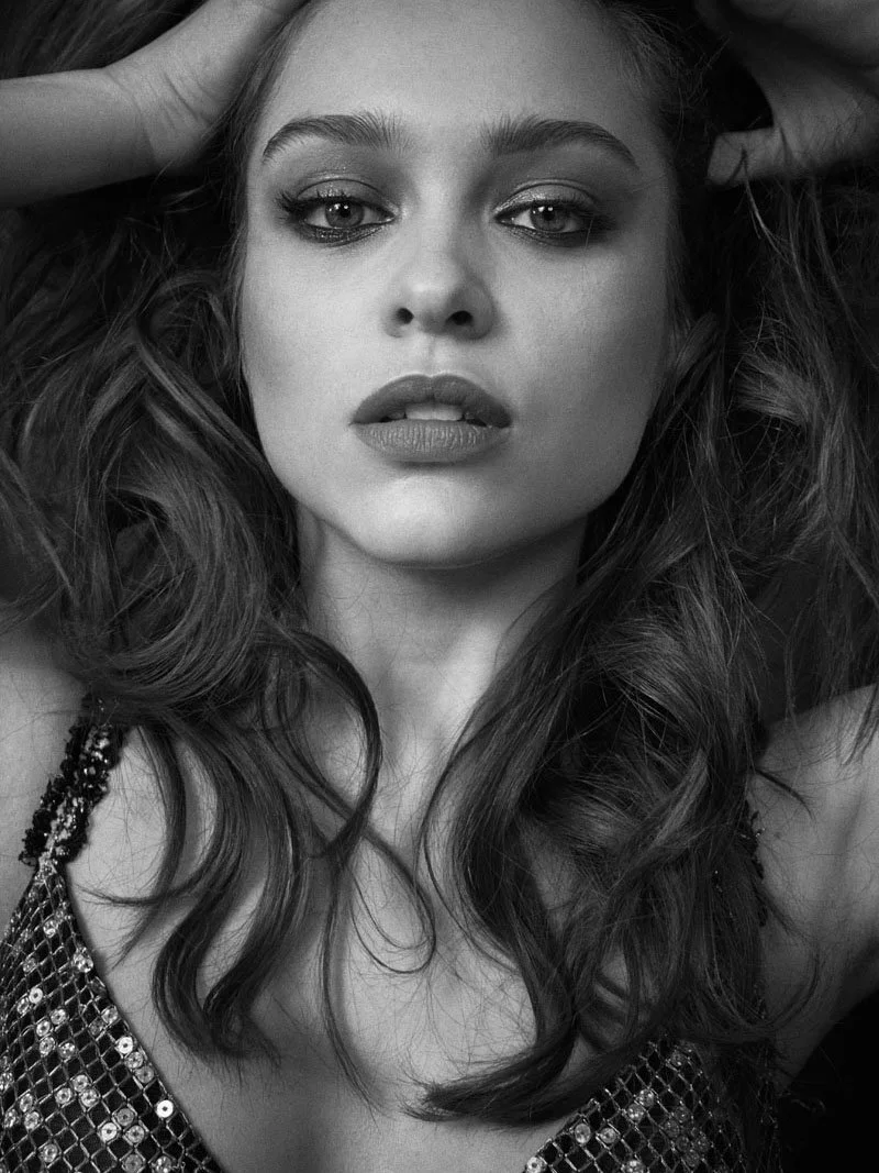 A black and white close-up photograph of a woman with long, wavy hair, looking directly at the camera with her hands in her hair.