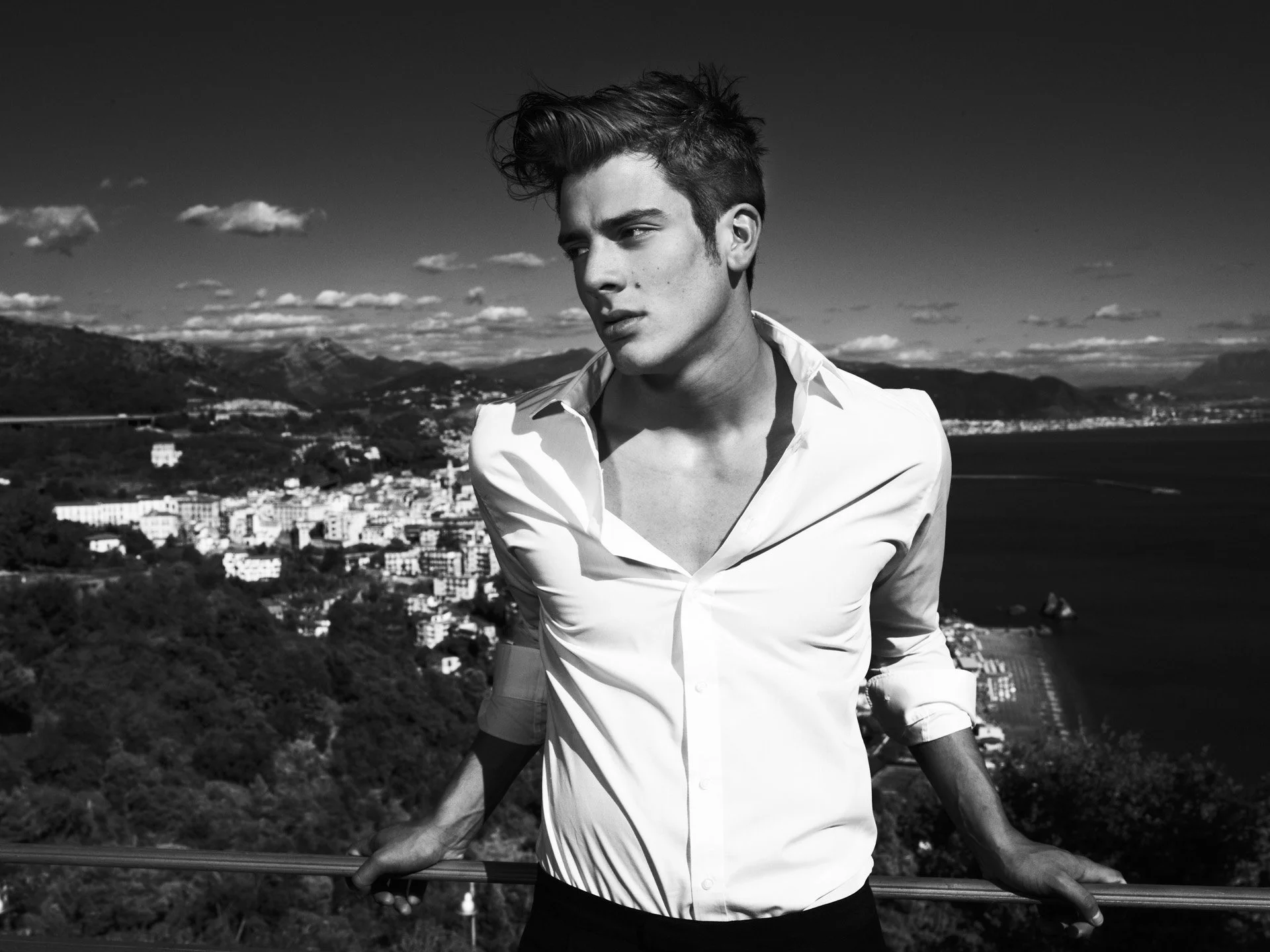 A black and white photo of a young man with styled hair and a serious expression, wearing a partially unbuttoned white shirt and standing outdoors with a cityscape, mountains, and a body of water in the background.