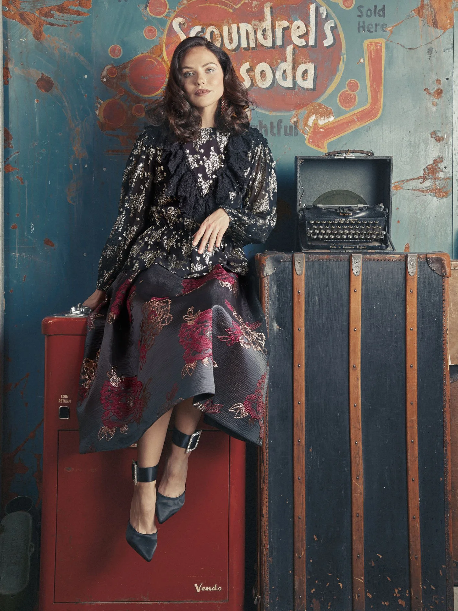 A woman with dark, wavy hair sitting next to a vintage typewriter in front of a weathered wall with a painted sign reading 'Soundrel's Soda.' She is wearing a black and silver floral dress with lace details, black ankle boots, and has her legs crosse