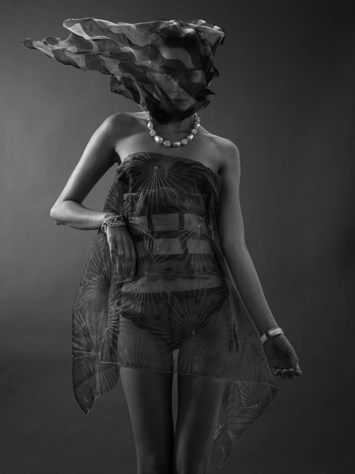 Black and white photo of a woman wearing a strapless dress with a pattern, layered with a sheer, patterned shawl or wrap. She has a large, distinctive headscarf or tulle wrap covering her hair, and is accessorized with a statement necklace, rings, br