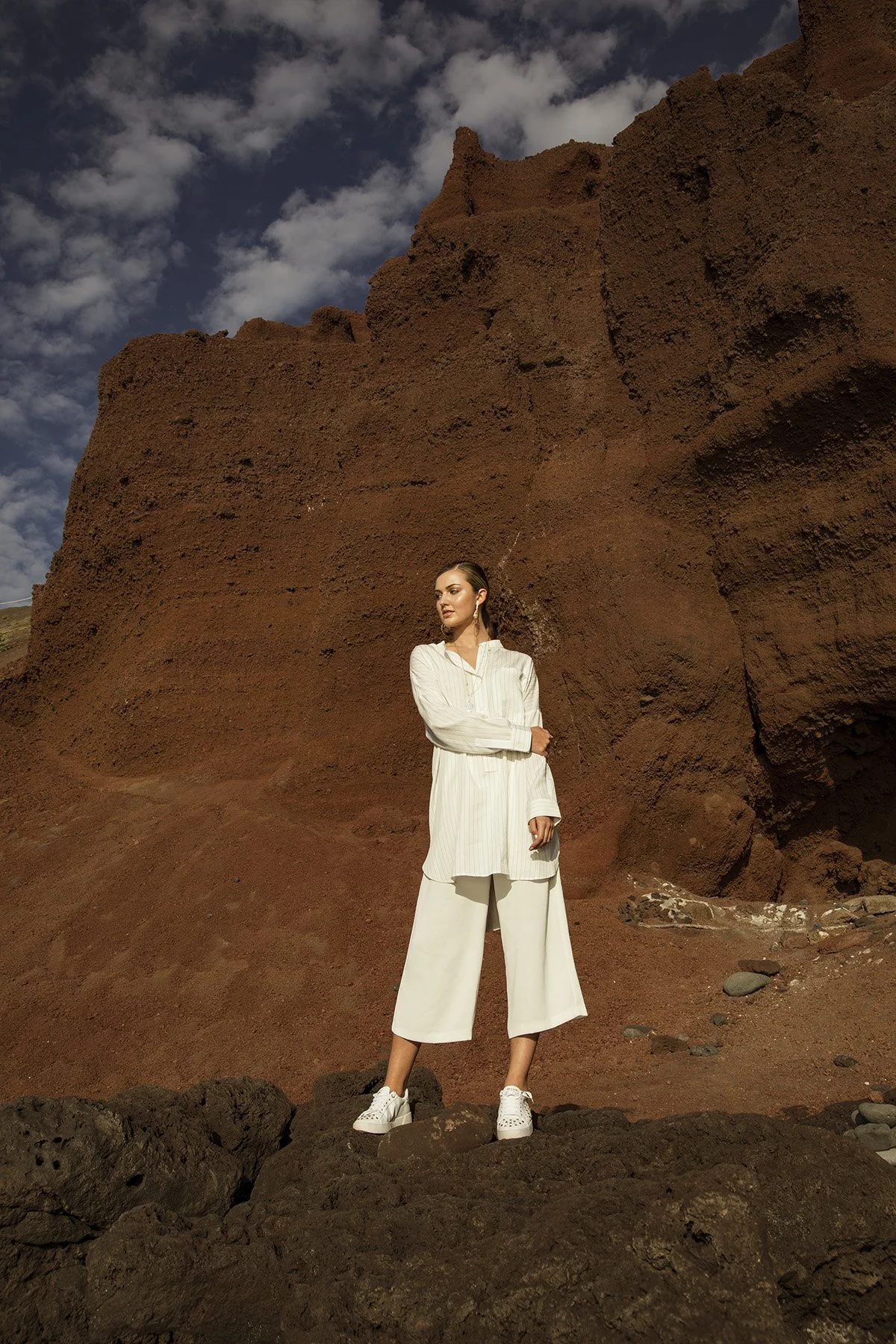 A woman in a white outfit standing on rocky terrain in front of a large reddish-brown rock formation under a partly cloudy sky.