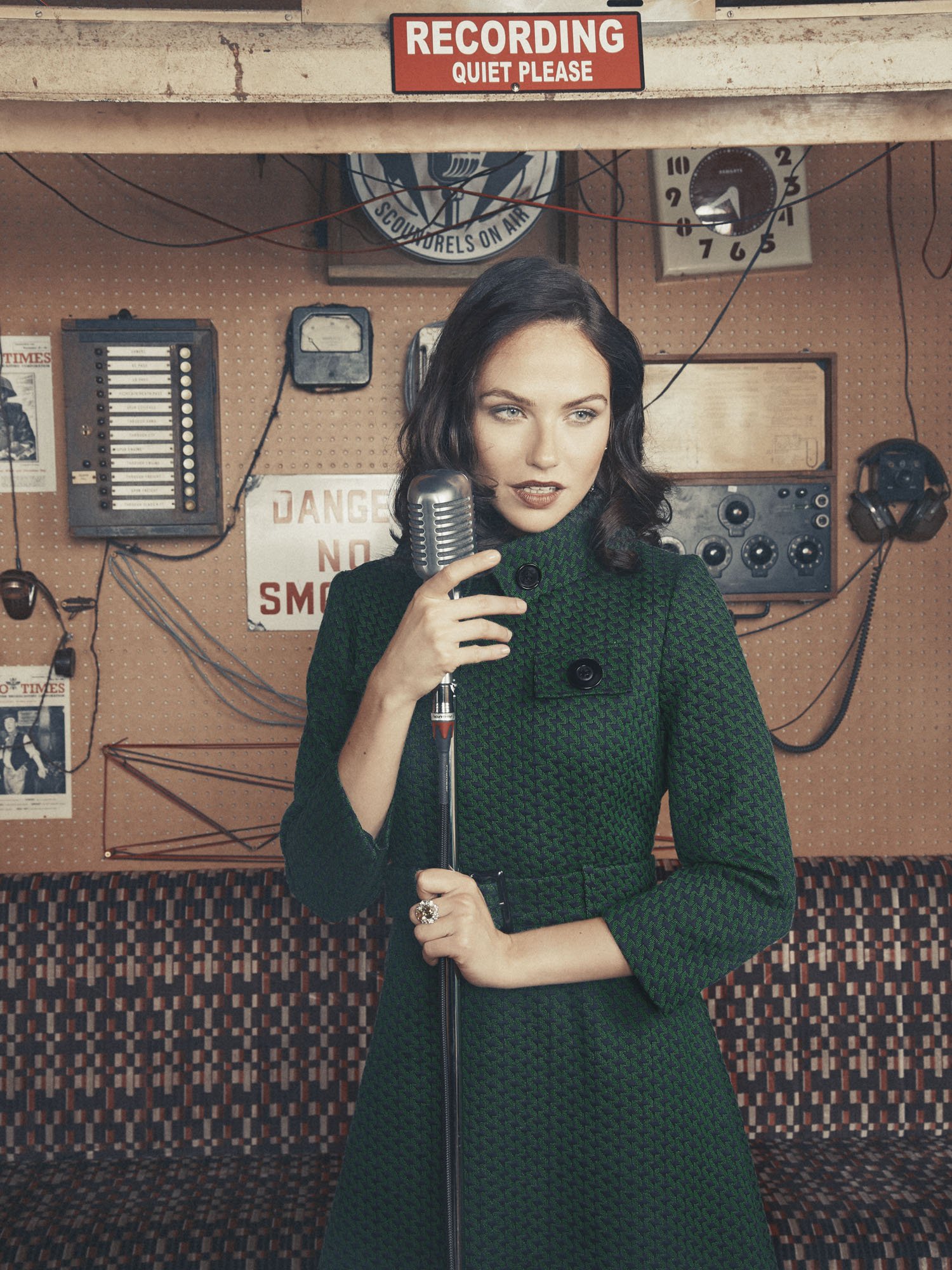 A woman in a green patterned coat holding a vintage microphone inside a recording studio, with various old radio and audio equipment in the background.