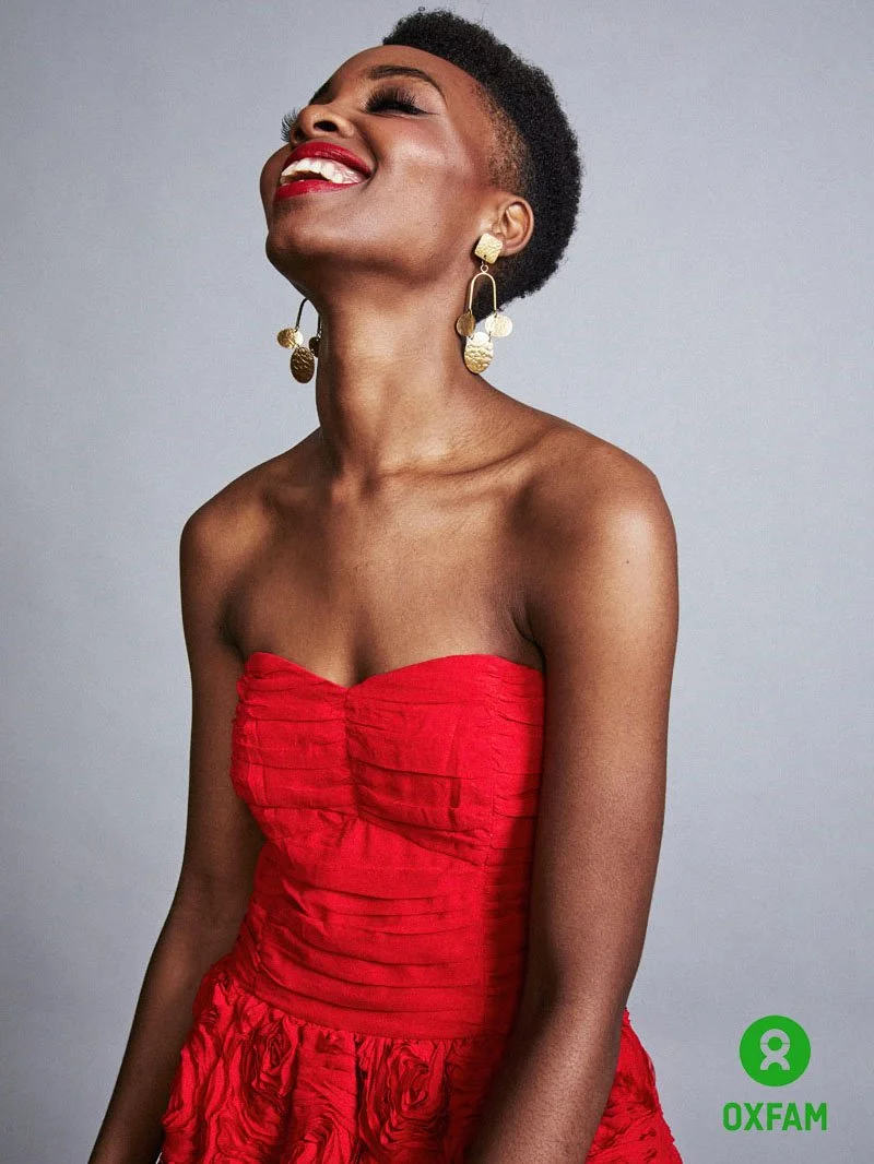 A woman with short curly hair wearing a strapless red dress and large gold earrings, smiling with eyes closed against a gray background, with an Oxfam logo in the bottom right corner.