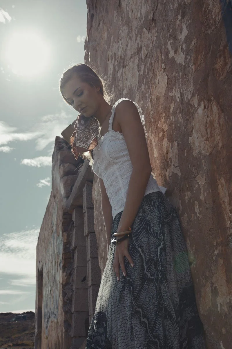 A woman standing outdoors against a textured stone wall, with the sun shining in the sky behind her.