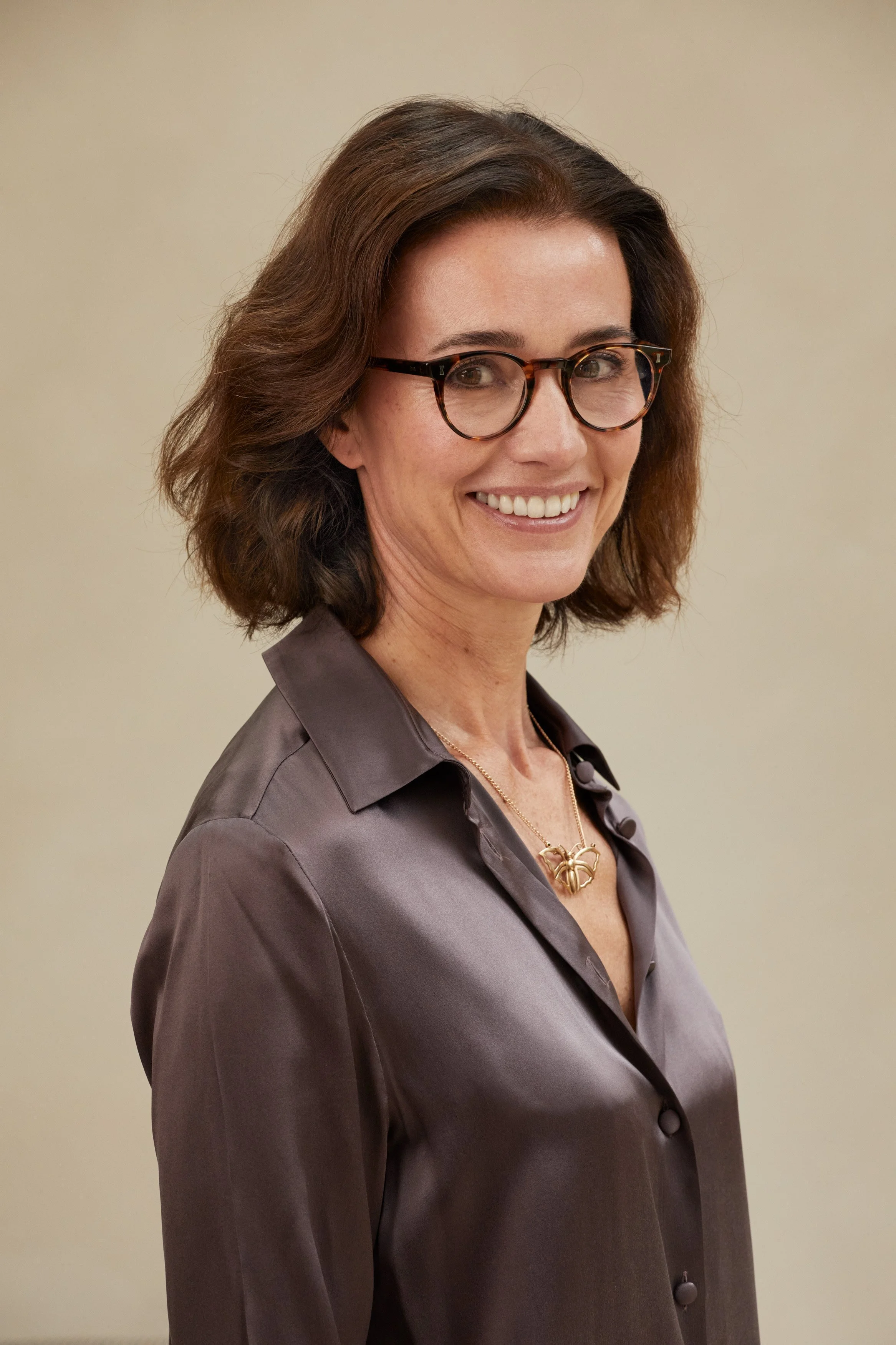 A woman with shoulder-length brown hair, wearing glasses, a gray satin blouse, and a gold necklace with a butterfly pendant, smiling in front of a plain beige wall.