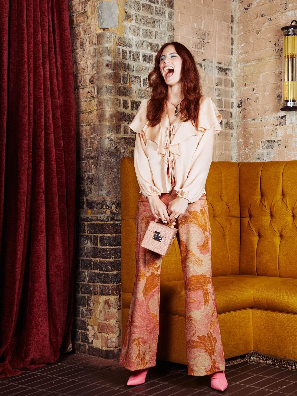 Woman laughing and posing in stylish vintage attire with a purse, standing in front of a brick wall and yellow sofa.