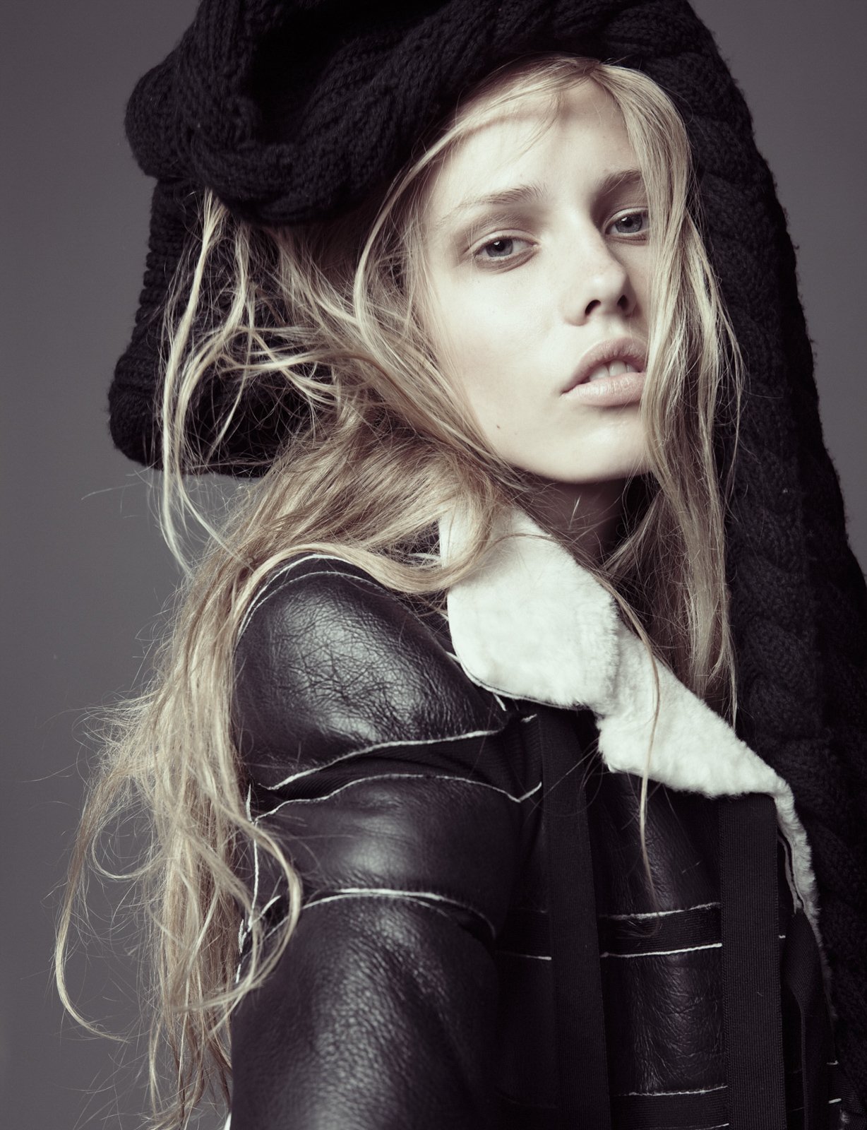 A young woman with long, wavy blonde hair wearing a black knit hat, a black leather jacket with a shearling collar, and a striped shirt under the jacket, looking into the camera with a neutral expression.