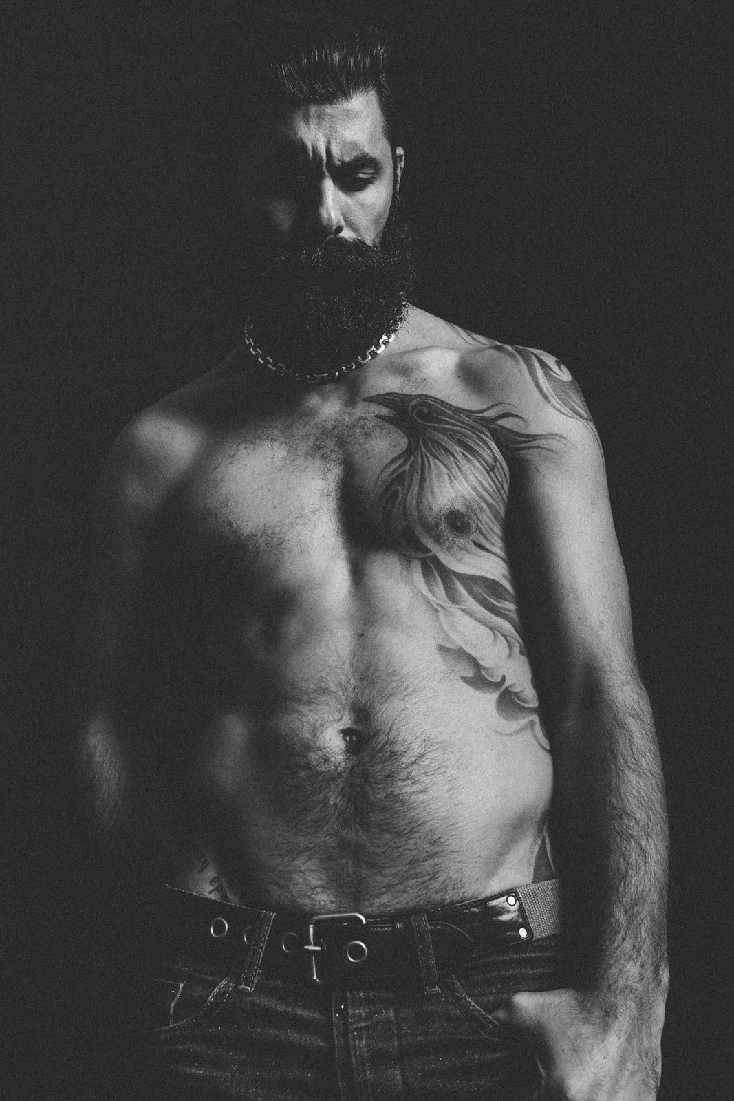 A black and white photo of a shirtless man with a beard and styled hair, wearing a chain necklace, with a large tattoo of a lion's head on his chest, standing against a dark background.