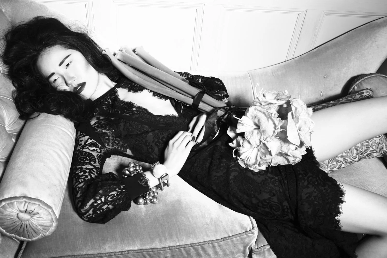 A woman with dark hair lying on a vintage-style sofa, wearing a black lace dress, holding a bouquet of flowers, with jewelry on her wrist and fingers, in a black and white photo.