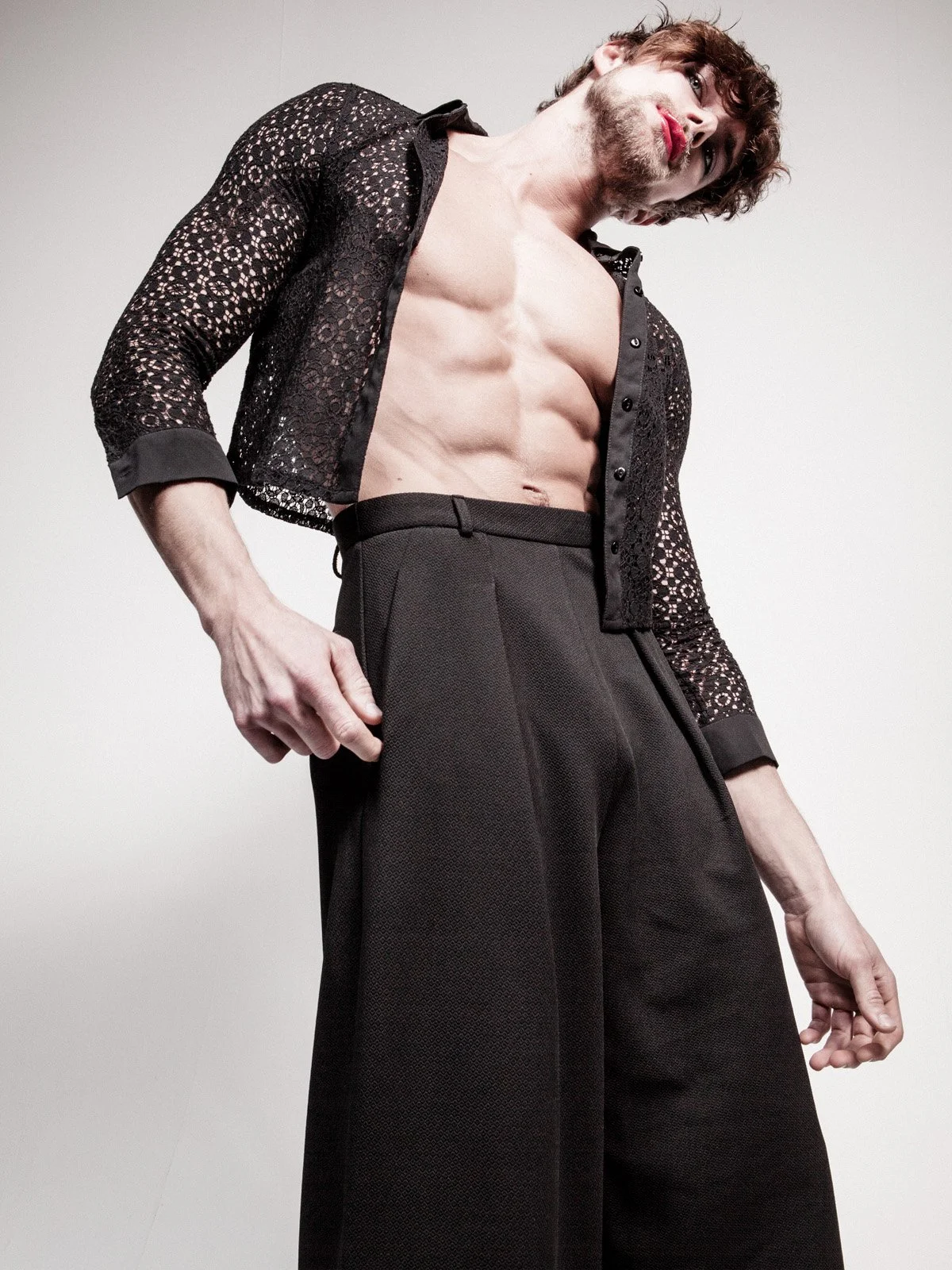 A shirtless man wearing black dress pants and a black lace blazer, showing well-defined abdominal muscles, stands against a plain gray background.