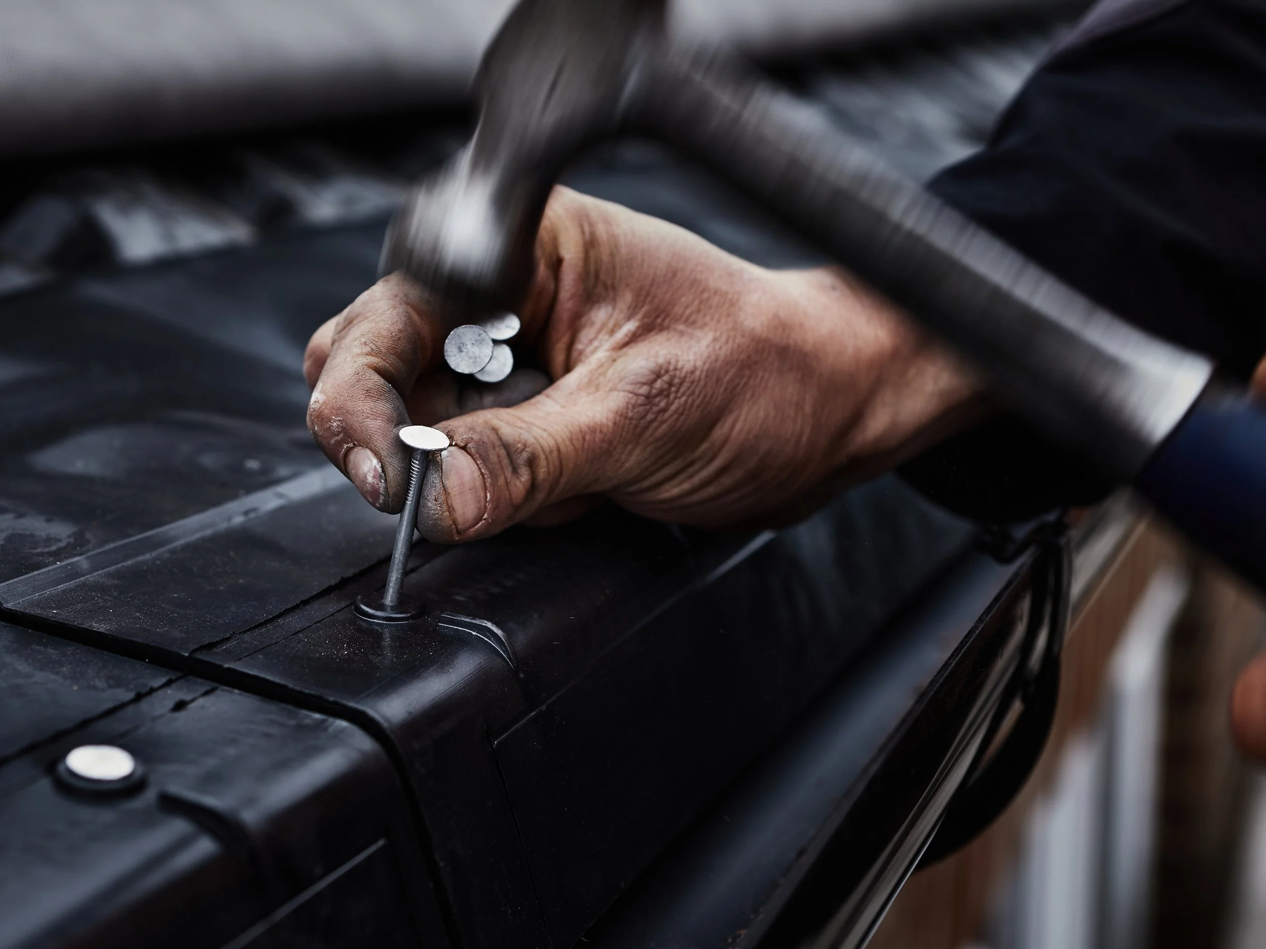 A person's hand using a hammer to hammer in a nail into a piece of black wood.