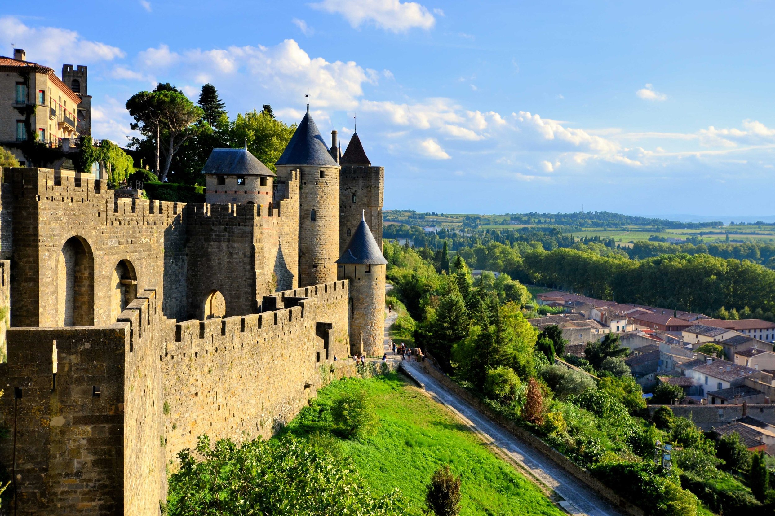 Scenic view of a historic stone castle with turrets and battlements, overlooking a lush green landscape and a small town under a bright blue sky with scattered clouds.