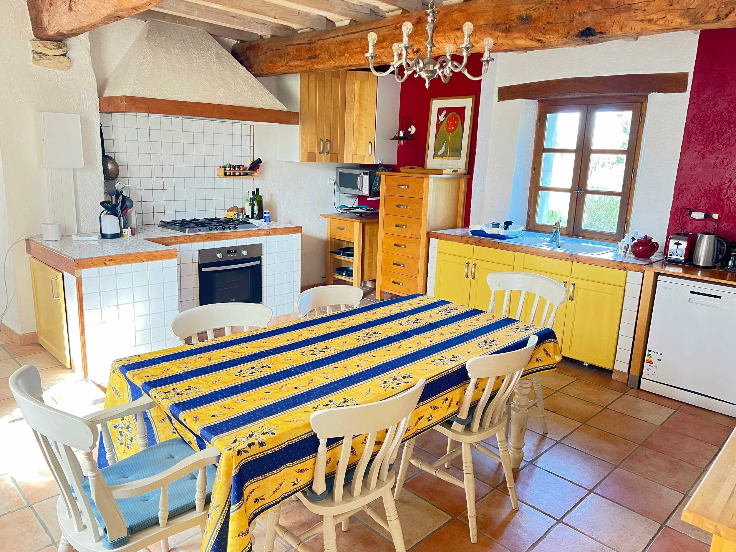 A cozy kitchen with yellow cabinets, a wooden dining table with a blue and yellow striped tablecloth, and white chairs. There is a window with wooden frames, a chandelier hanging from exposed wooden beams, and various kitchen appliances and decor on the countertops.