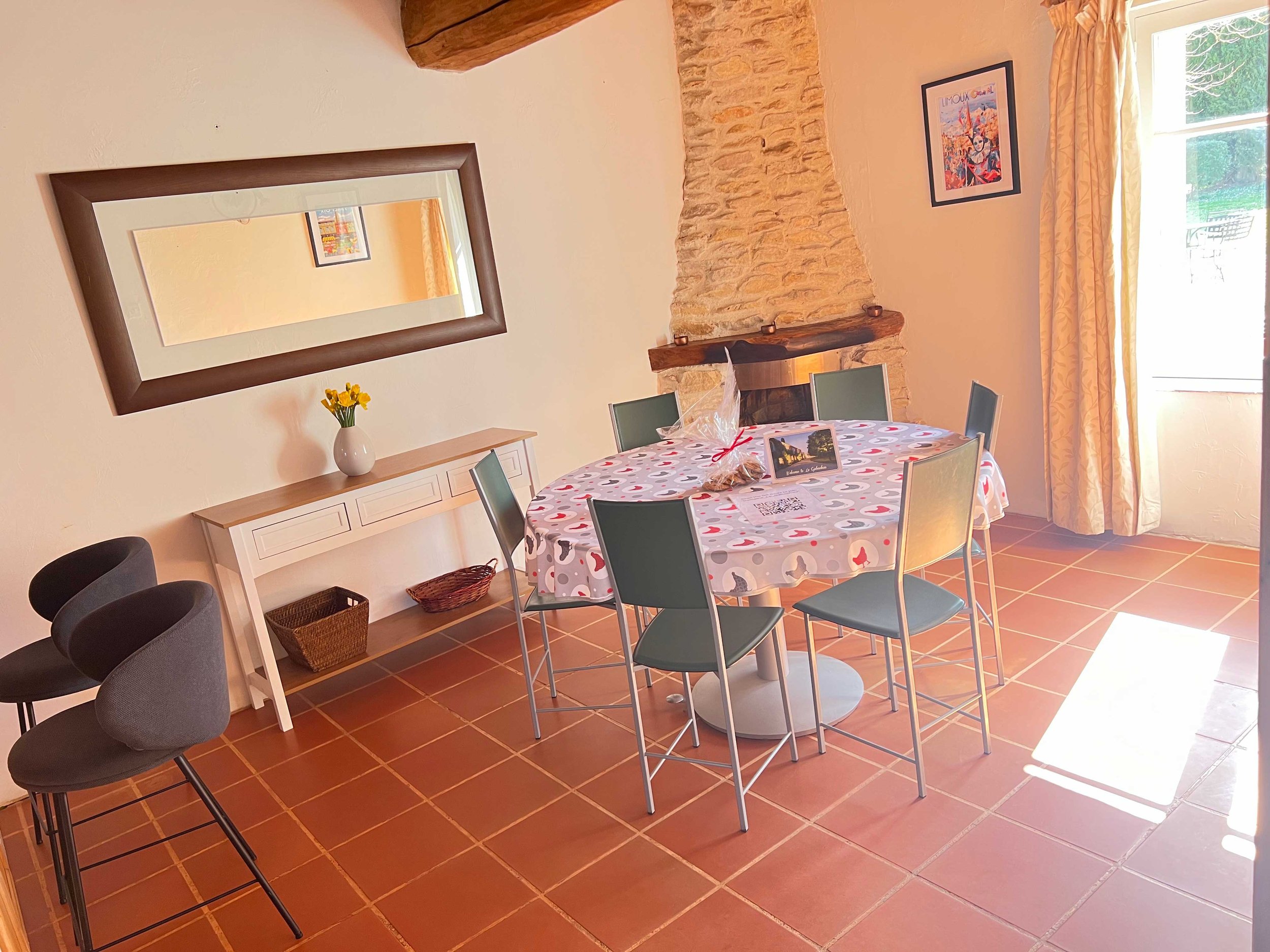 A cozy dining room with a round table covered in a heart-patterned tablecloth, surrounded by five chairs. There is a kitchen counter with a vase of yellow flowers, a mirror on the wall, a brick fireplace with candles, and framed artwork. Large windows with yellow curtains let in natural light.