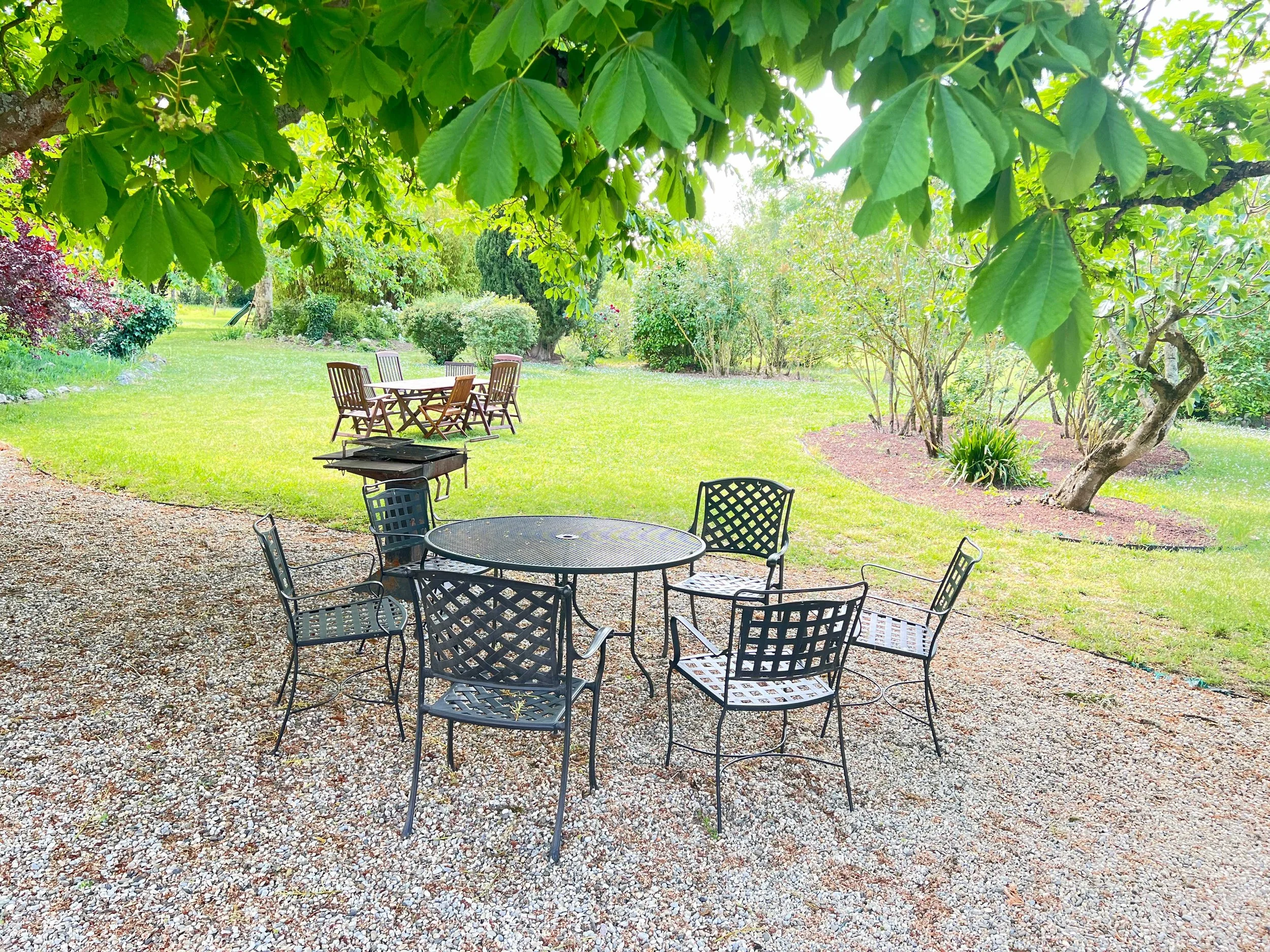 Outdoor patio area with a round metal table and black metal chairs, surrounded by a lush green garden with trees, shrubs, and a grassy lawn.