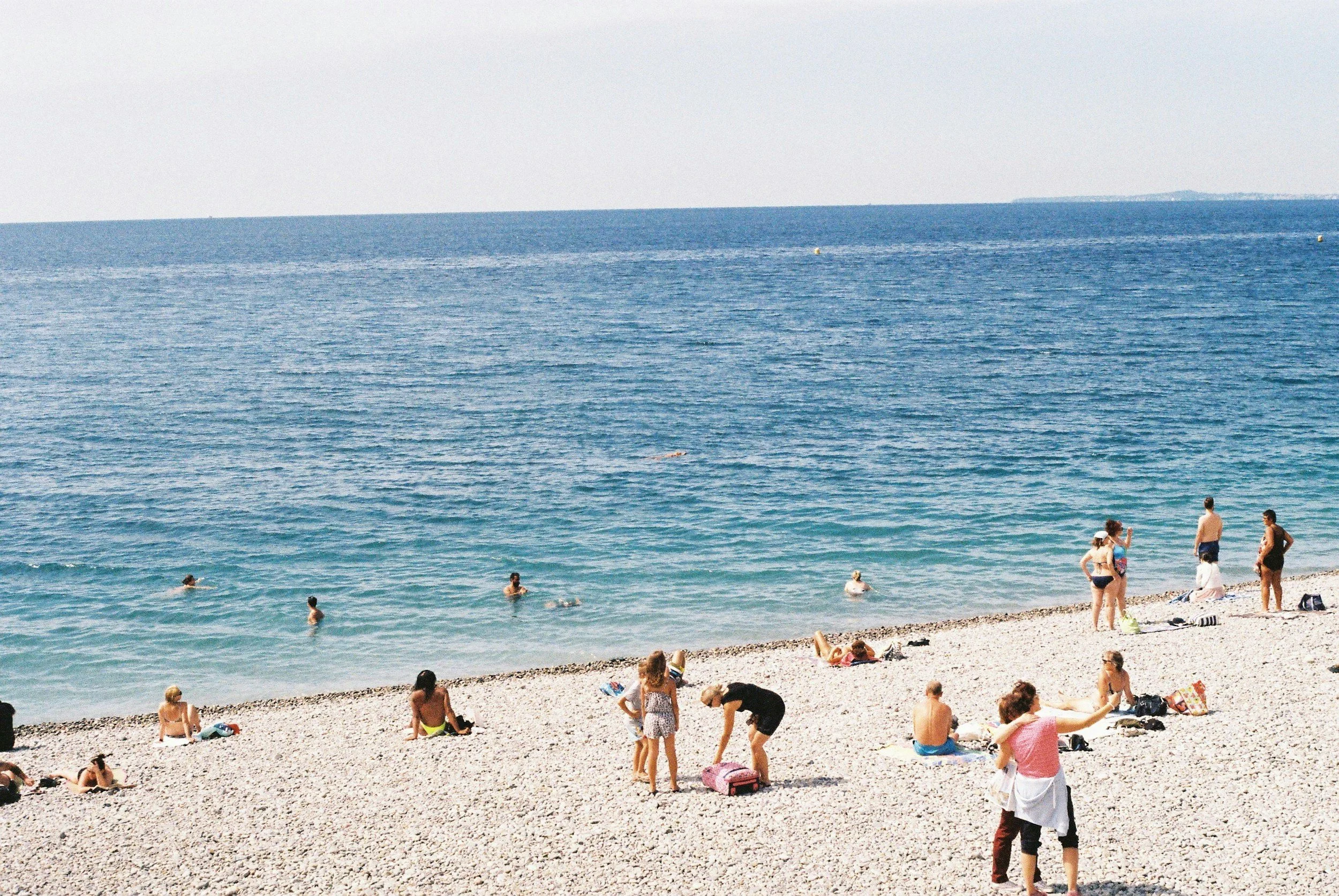 People enjoying a day at the beach with the ocean in the background, some swimming in the water, others sitting or standing on the sandy shore.