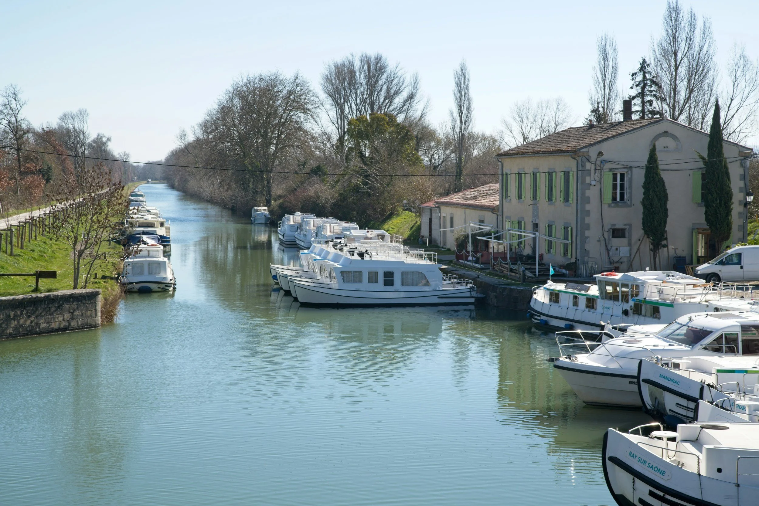 A narrow canal with mainly white boats moored along the right side. A large old building with green window shutters is on the right. Bare trees line the canal and extend into the background, with a clear blue sky overhead.