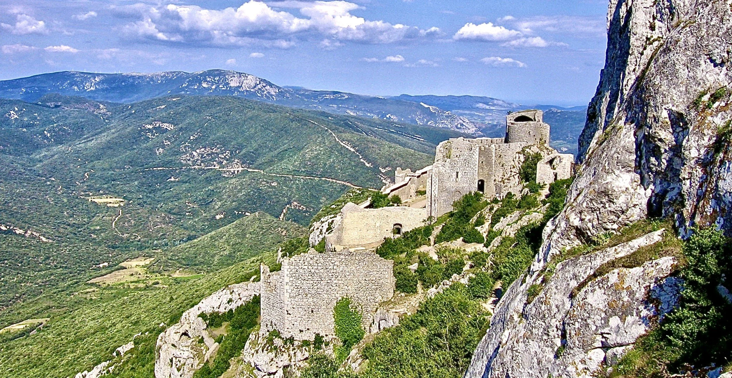 A scenic view of an ancient stone castle built on a mountain cliff overlooking lush green hills and distant mountains under a partly cloudy sky.