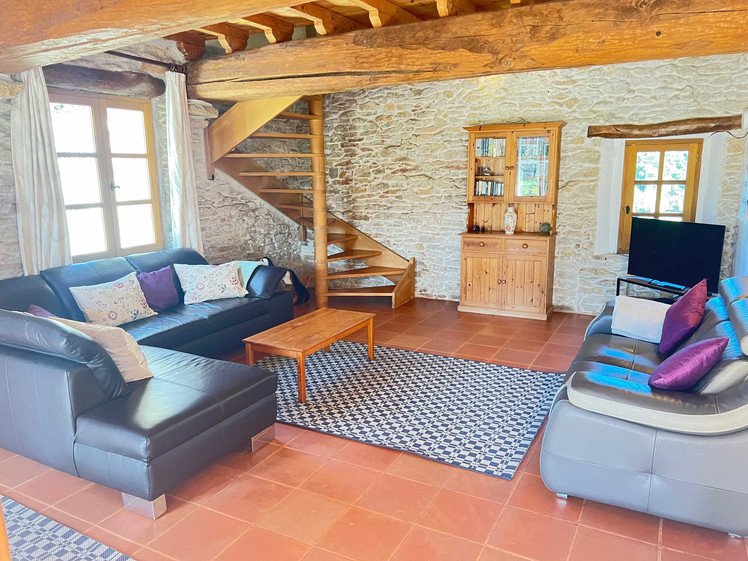 A cozy living room with stone walls and wooden beams. Contains two leather sofas with pillows, a wooden coffee table, a bookshelf, and a television. Natural light comes through windows with white curtains.