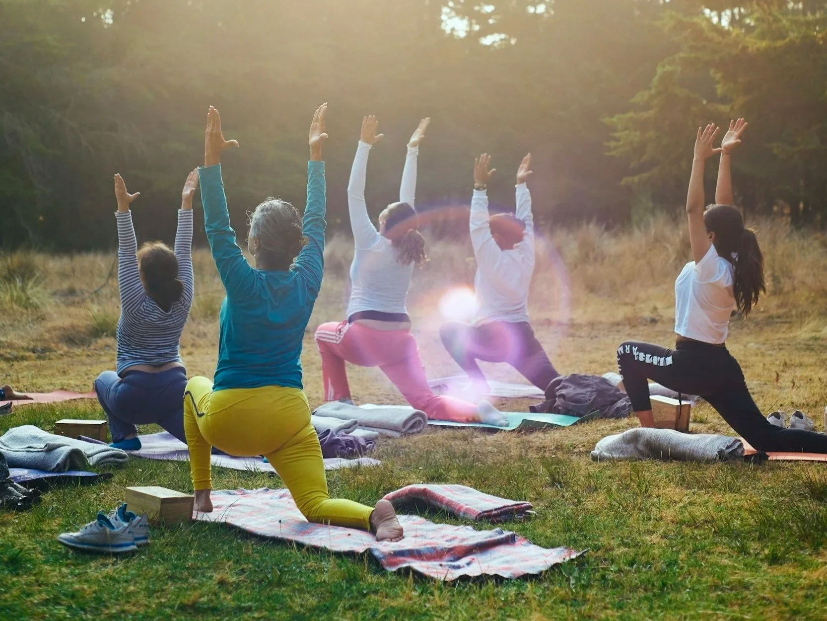 Group of seven women practicing yoga outdoors on mats during sunrise or sunset, with trees in the background.