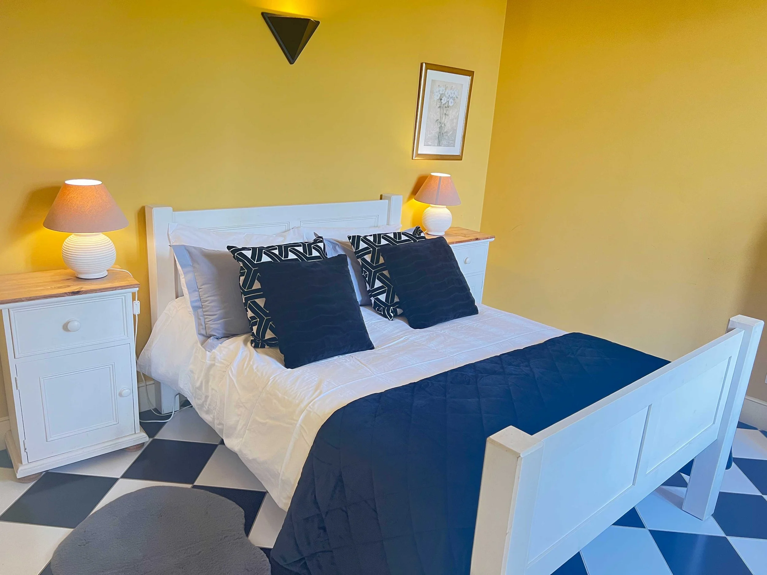 A bedroom with yellow walls, a white bed with black and white geometric pillows and a black bedspread. Two white nightstands with large white ceramic lamps with pink shades. A framed floral artwork hangs on the wall above the bed, and a black triangle wall decoration is above it. The floor has black and white checkered tiles and a large dark gray area rug at the foot of the bed.