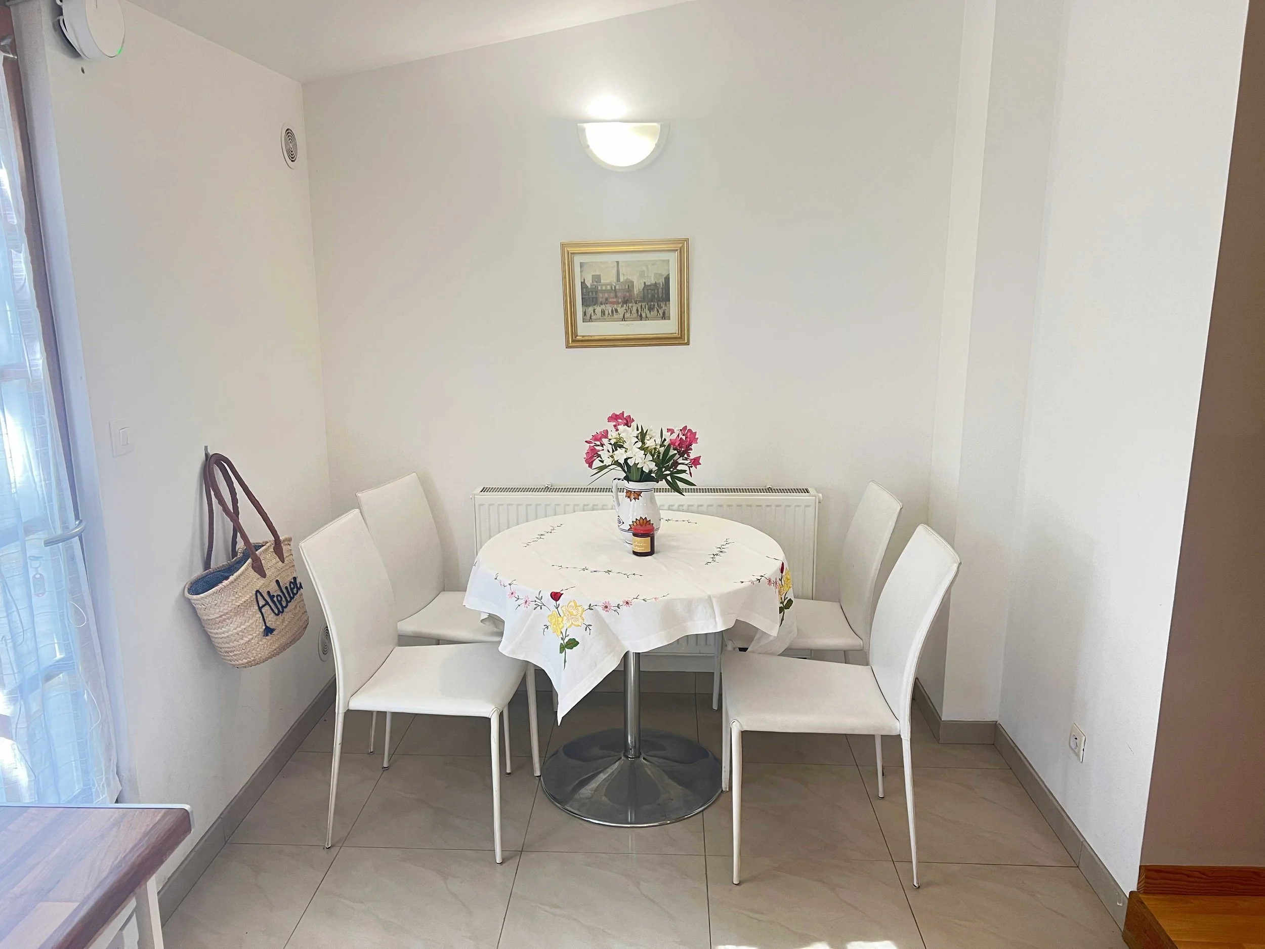 Dining area with a round table covered by a floral tablecloth, surrounded by six white chairs, a vase with pink and white flowers on the table, a framed picture on the wall, and a wicker bag hanging on a hook.