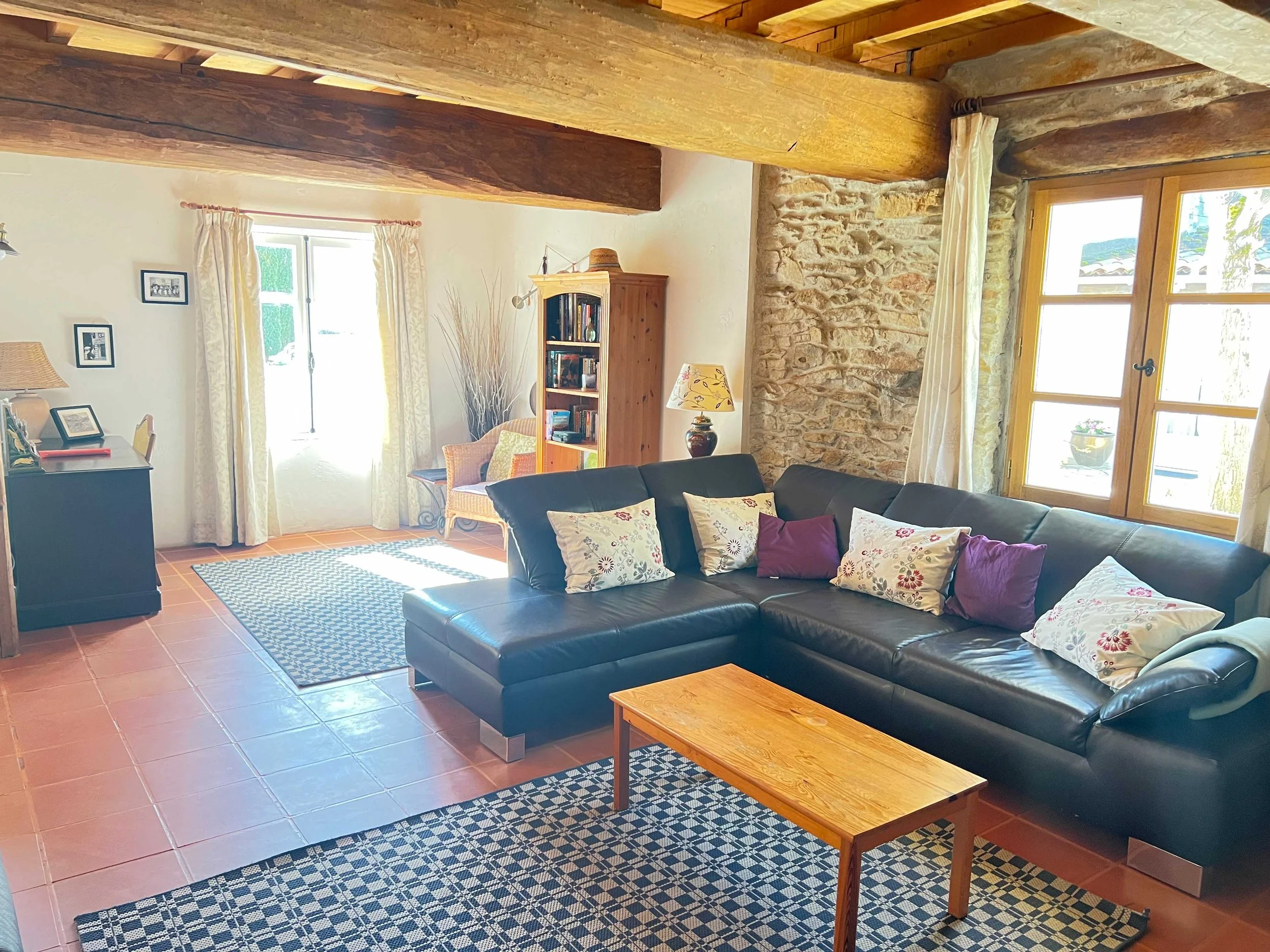 Living room with leather sofa, wooden coffee table, bookshelf, framed photos, and windows with curtains letting in natural light.