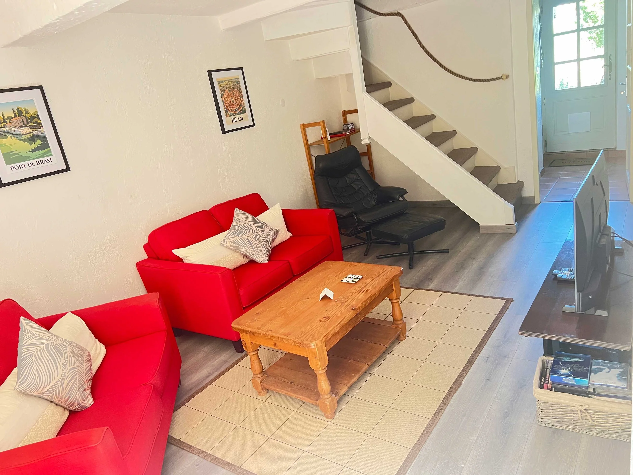 Living room with red sofa, beige pillows, wooden coffee table, black lounge chair, staircase with gray carpet, and framed posters on white wall.