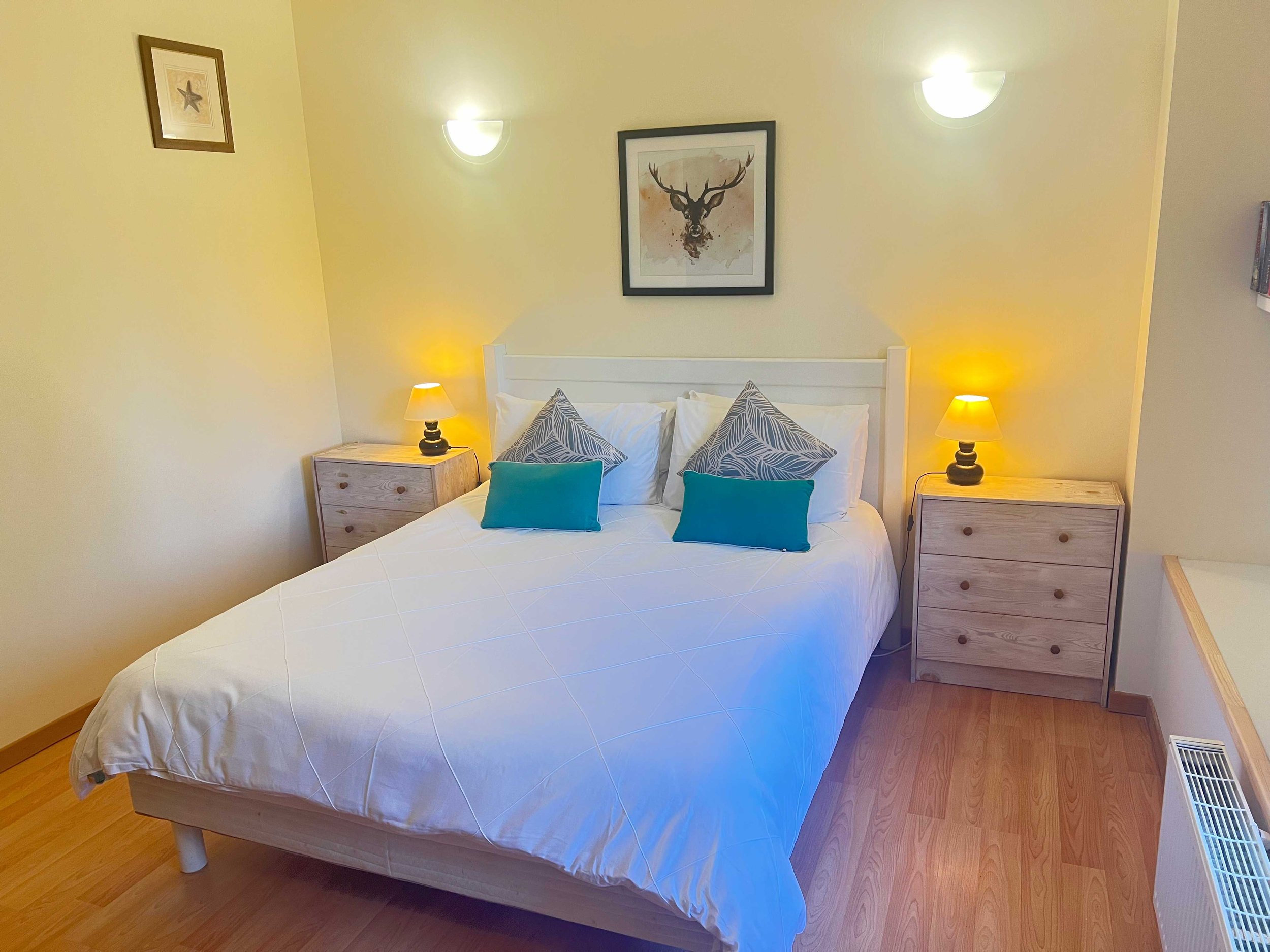 A bedroom with a bed having white linens and decorative pillows, two wooden nightstands with small lamps, yellow painted walls, framed art of a starfish and a deer head, hardwood floor, and a heater vent.