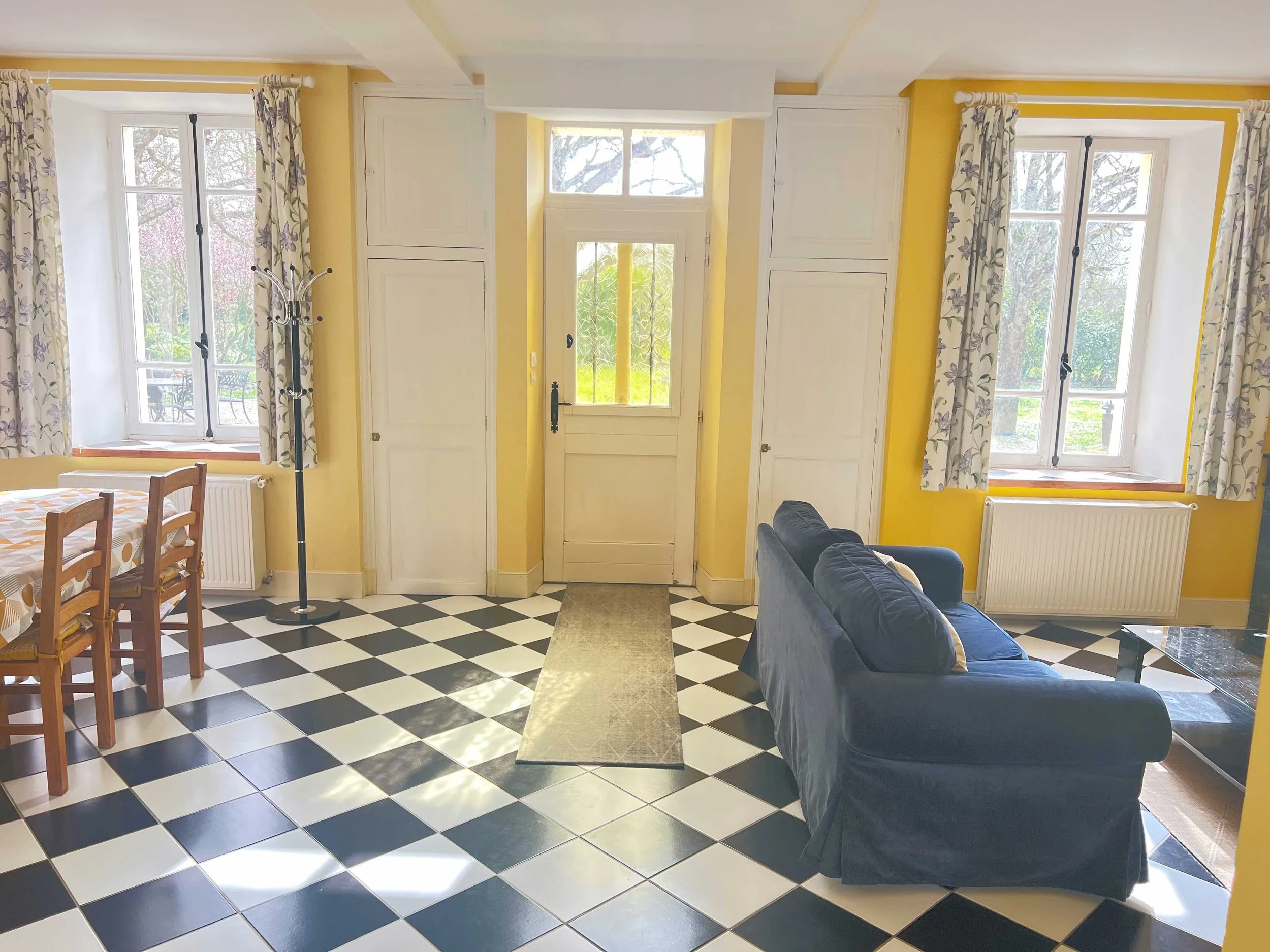 Living room with yellow walls, two large windows with floral curtains, a front door with a window, a blue sofa, a wooden dining table with chairs, and a black-and-white checkered floor.