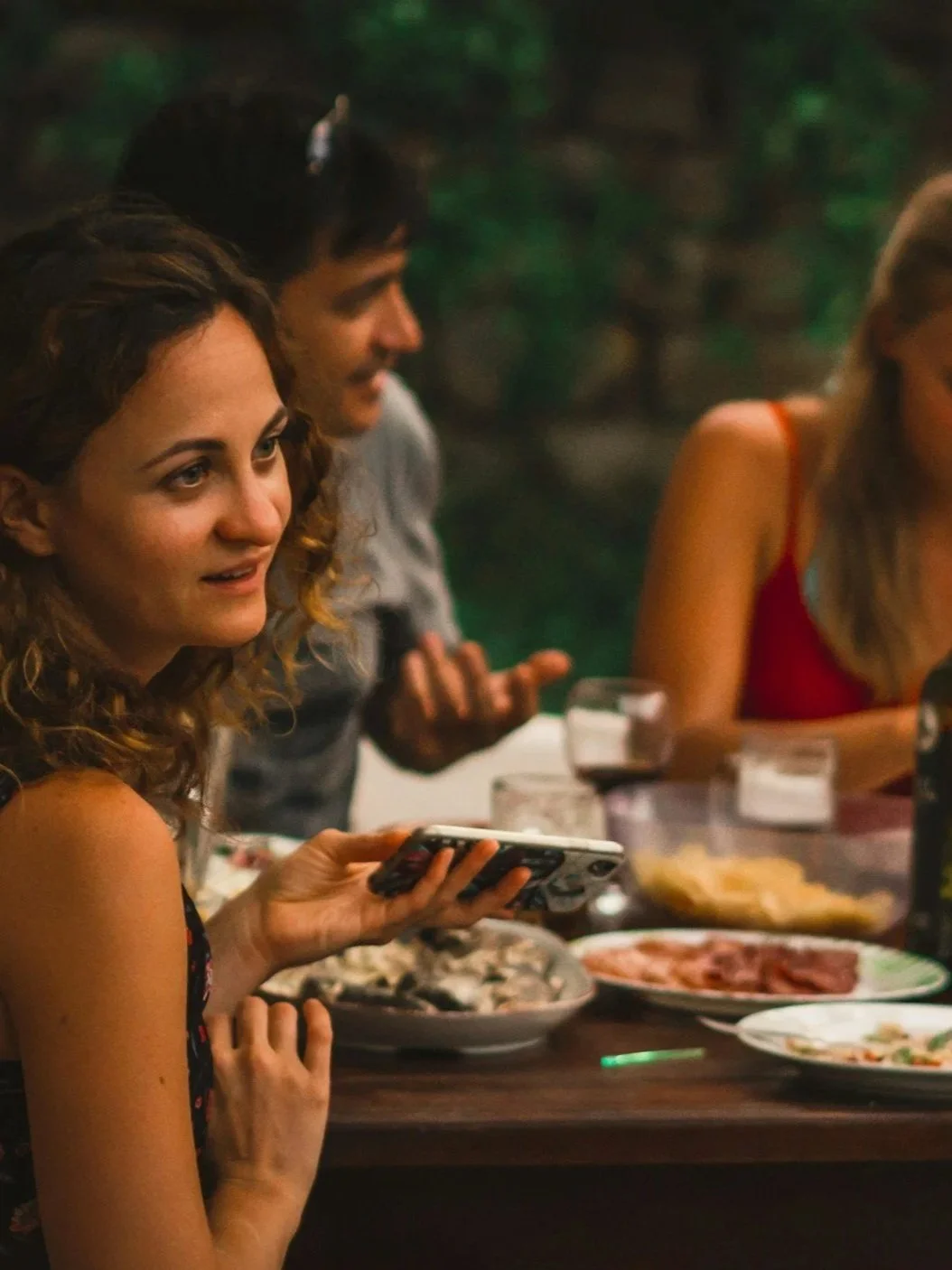 People sitting at a dinner table with food including meat and vegetables, one person holding a smartphone, in a dimly lit setting.
