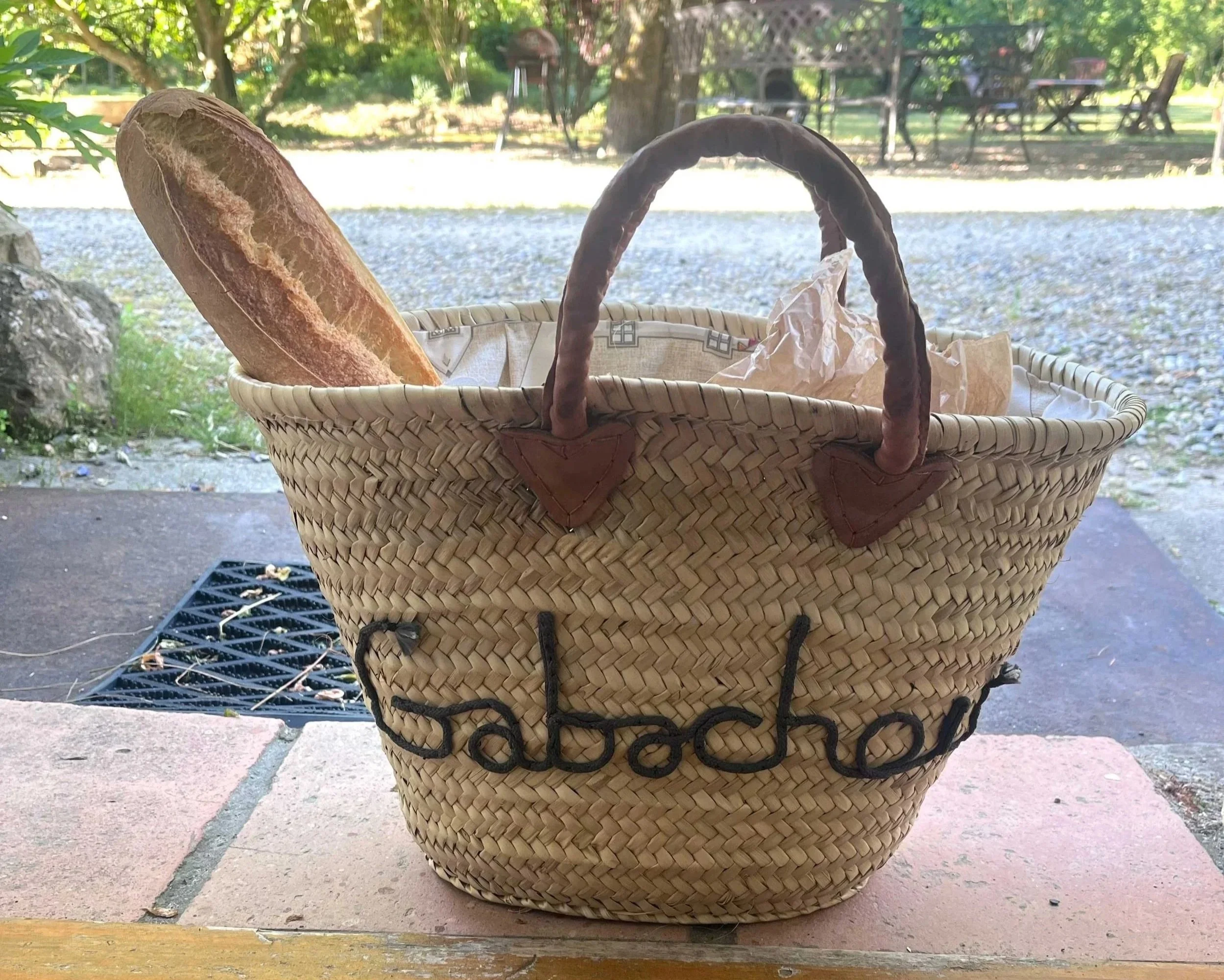 A wicker basket with a handle containing a loaf of bread wrapped in paper, set outdoors on a table with trees and a gravel yard in the background.