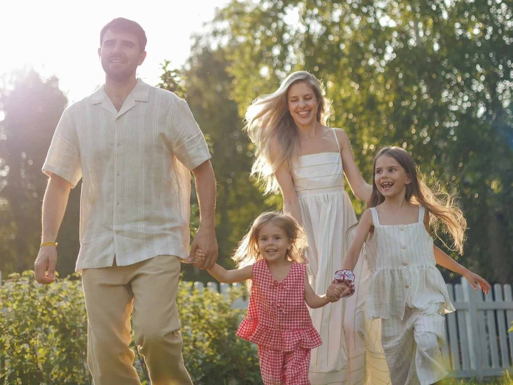 A family of five, including two young girls, a woman, and a man, walking hand-in-hand in a sunny outdoor setting with trees and a white fence in the background.