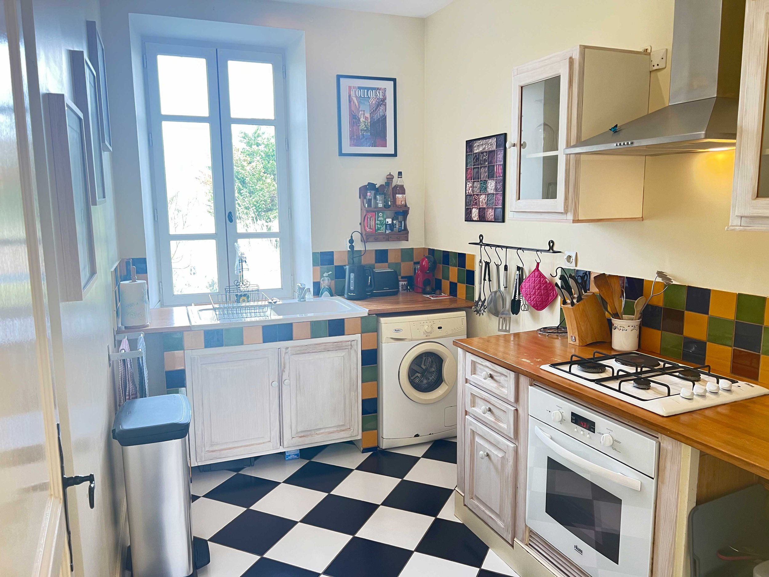 A compact kitchen with white cabinets, colorful tile backsplash, and black and white checkered floor, featuring a window, small shelf with spices, and various kitchen appliances and utensils.