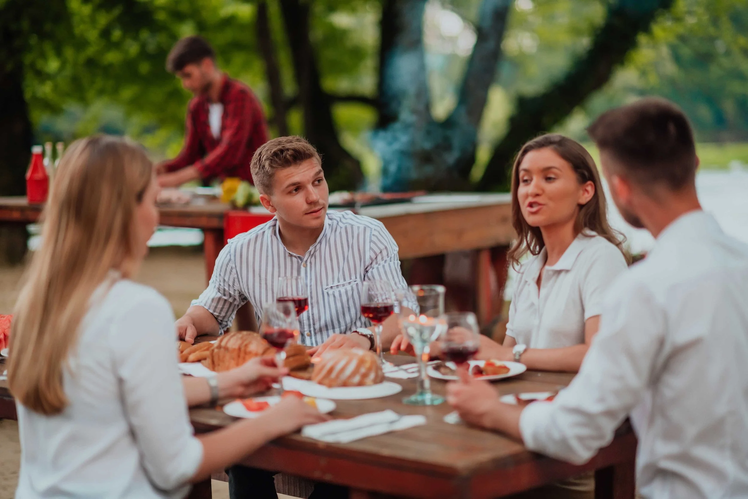 People enjoying a meal outdoors with a table set with food and drinks, surrounded by green trees.