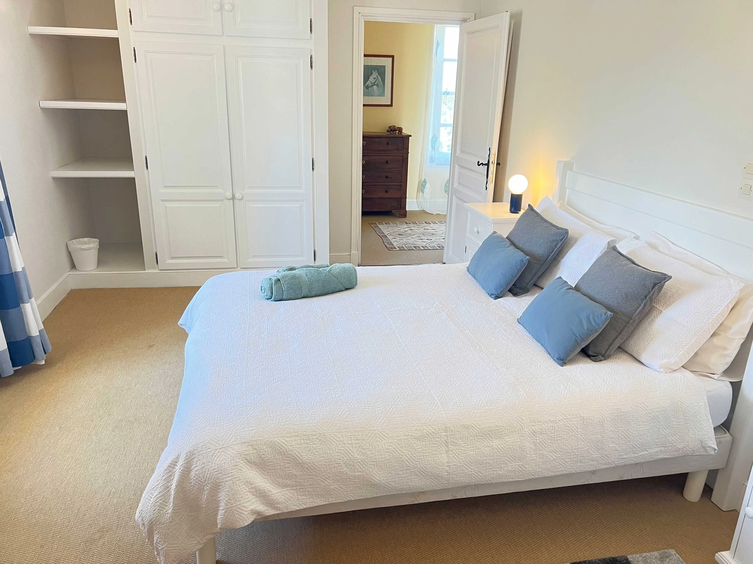 A neatly made bedroom with a white bed, blue and gray pillows, a nightstand with a round lamp, and a closet. An open door reveals a hallway with a wooden dresser and a framed picture of a horse.