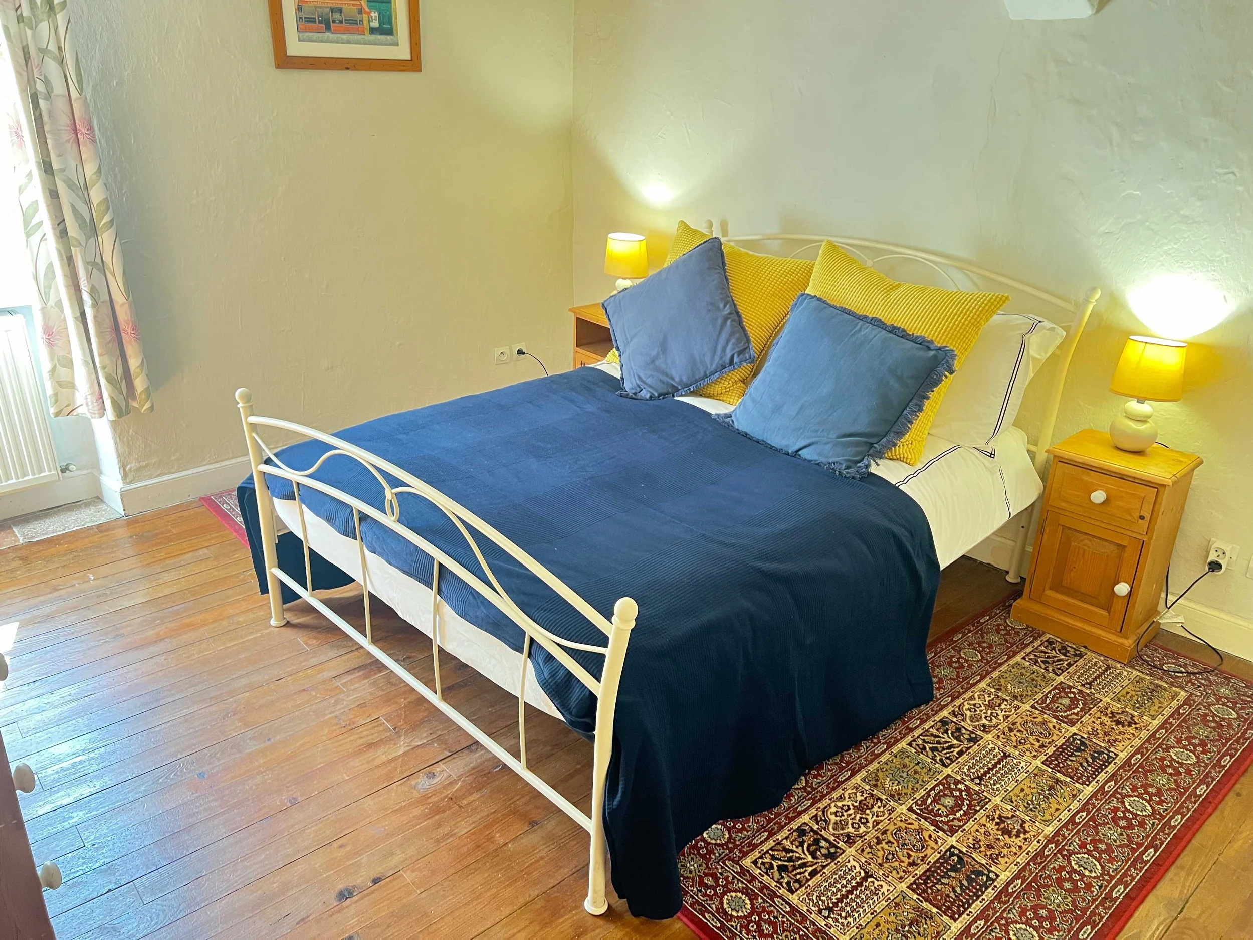 A bedroom with a white metal bed frame, blue and yellow bedding, yellow lamps, a wooden nightstand, framed artwork on the wall, a patterned rug, and hardwood floors.
