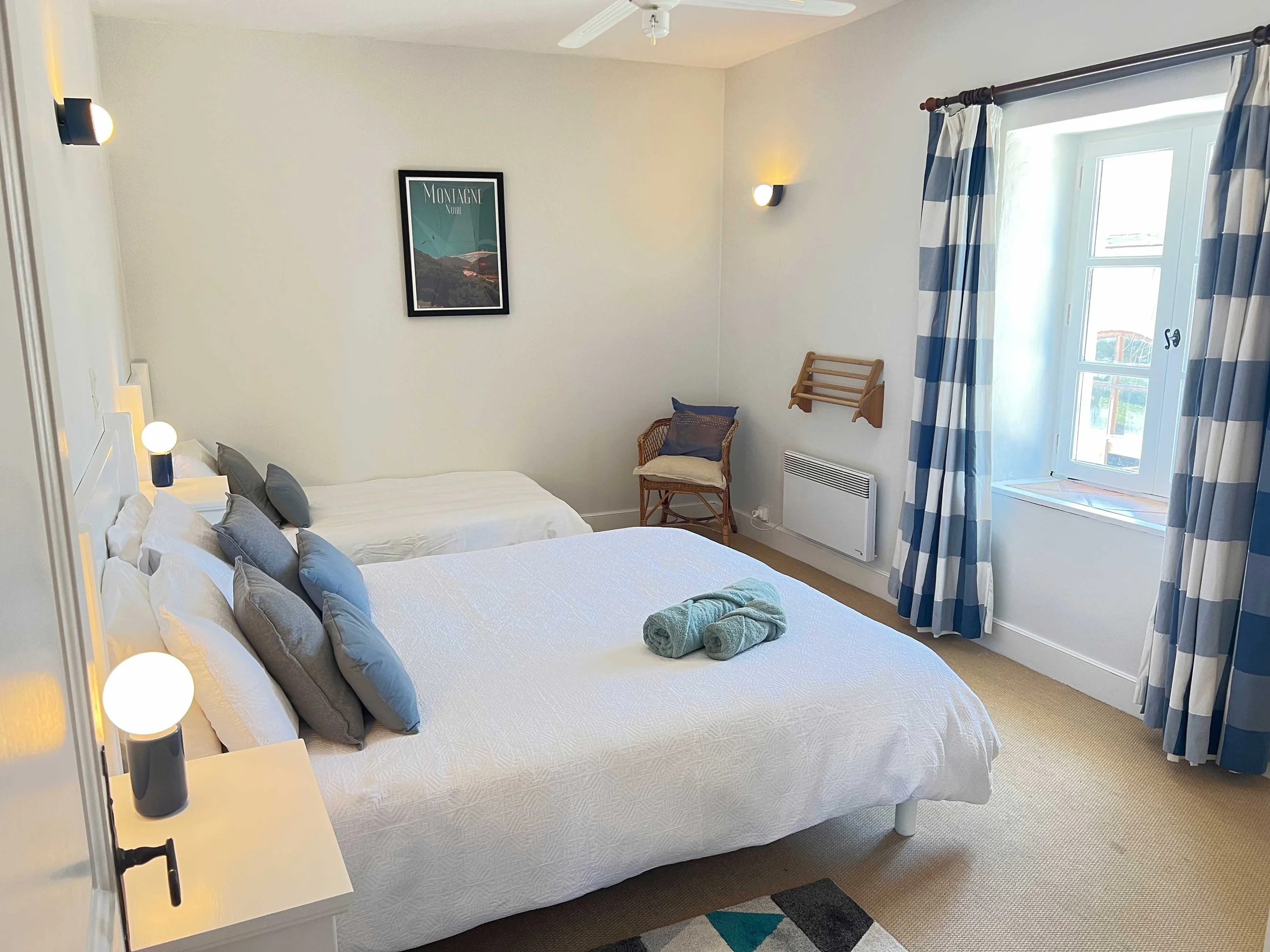 A bedroom with a white bed, multiple gray pillows, and two rolled towels on it. To the right there are blue and white checkered curtains and a window. There are wall-mounted lights and a picture of Montagne Noir on the wall, with a wicker chair and a small wooden rack beneath it.