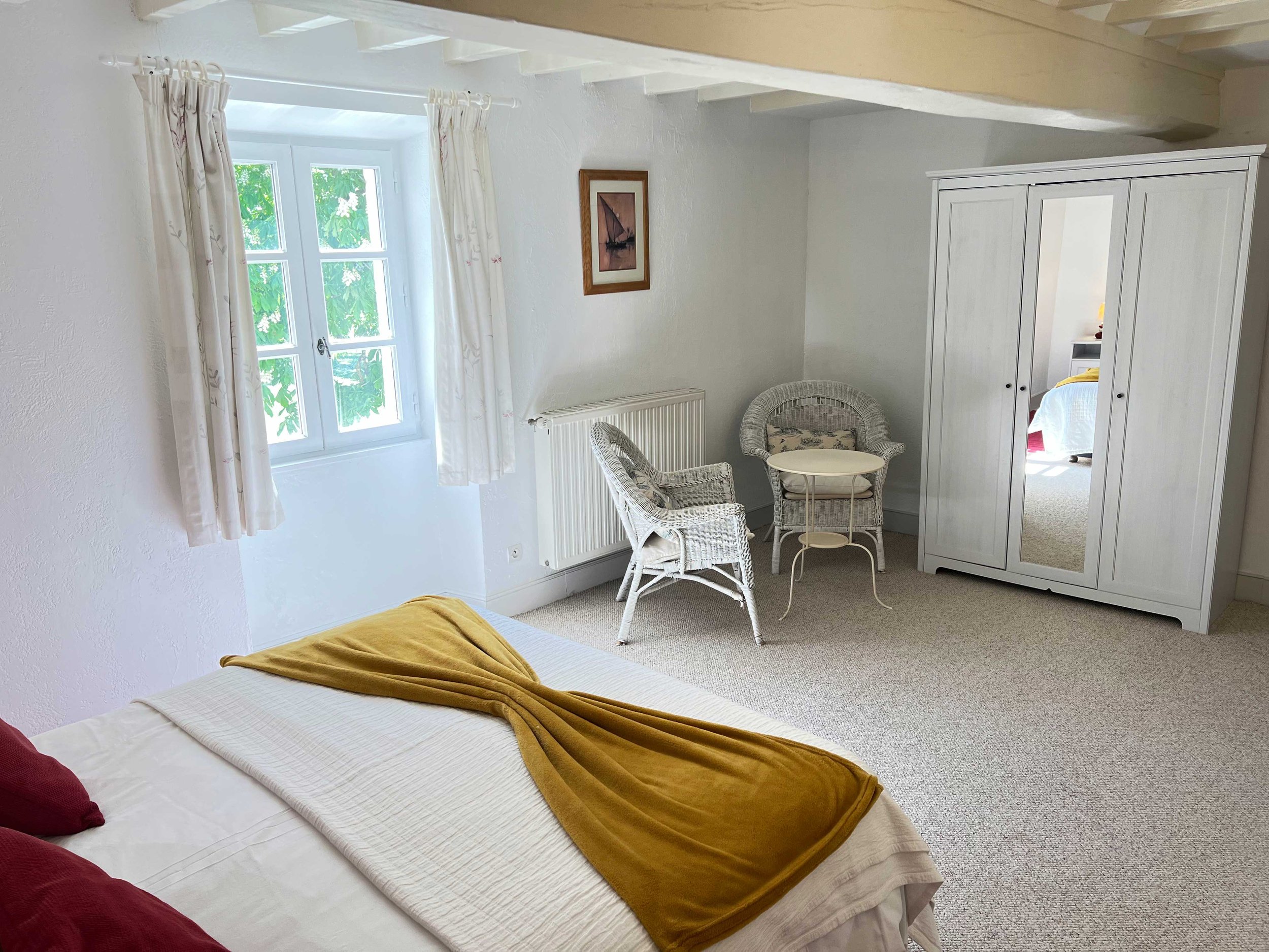 A bedroom with a bed in the foreground covered with a white sheet and a mustard-colored blanket, a window with white curtains, a small round table with wicker chairs, a white wardrobe with a mirror, a framed picture of a sailboat on the wall, and a door leading to another room.