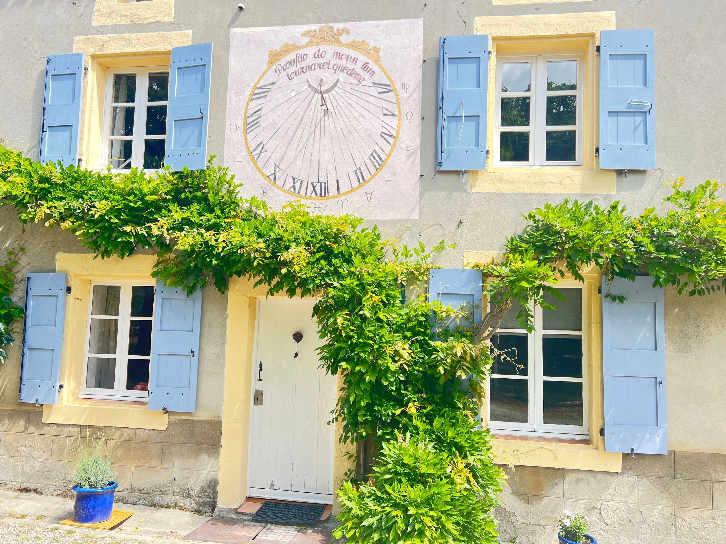A house with light yellow walls, blue window shutters, and a white door. There is a large clock painted on the exterior wall with Roman numerals and a phrase in French. Green bushes and potted plants decorate the front yard.