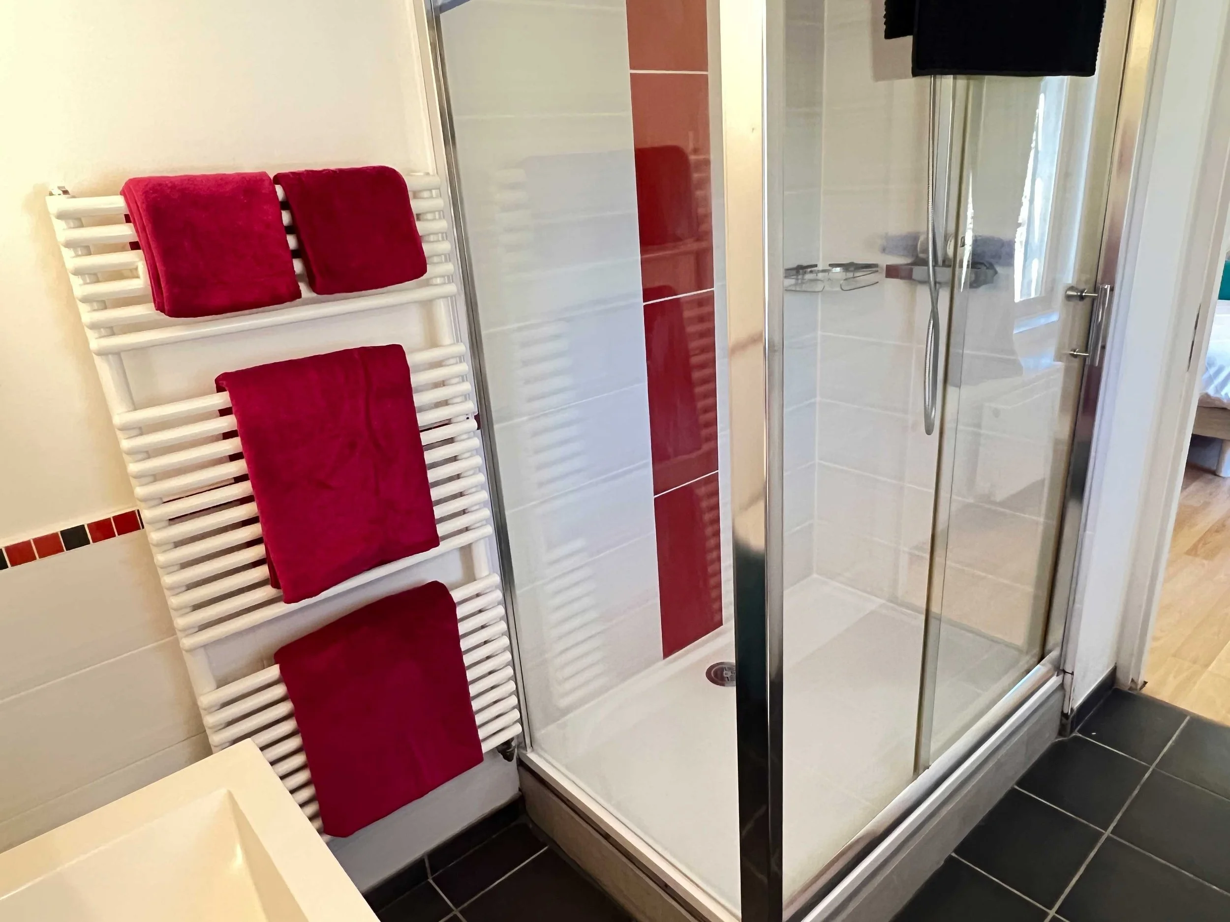 A bathroom shower enclosure with white tiles, red decorative tiles, a metal showerhead, and a glass door. Adjacent to the shower, there is a wall-mounted towel radiator with three red towels hanging on it.