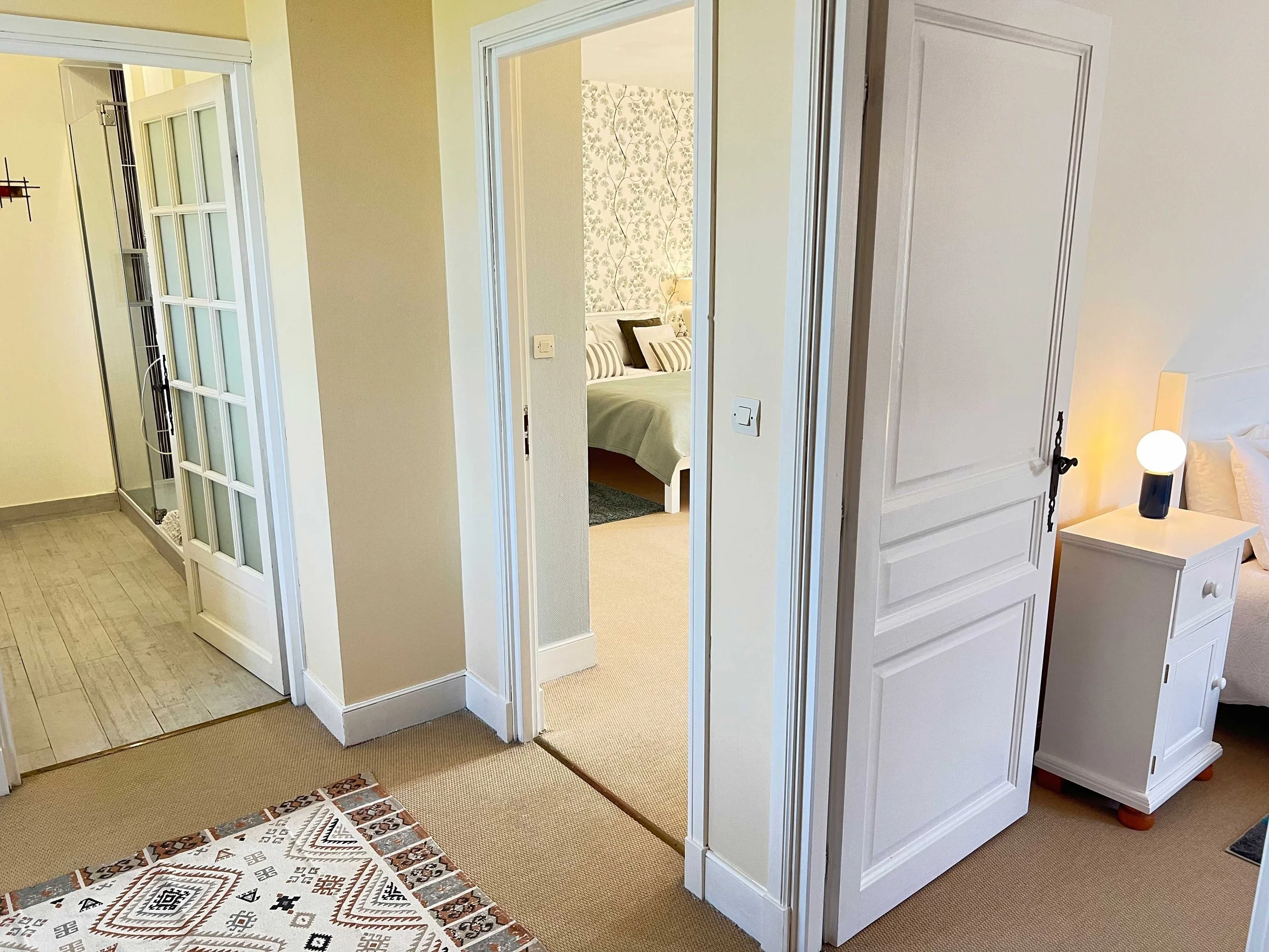 View of a hallway leading into a bedroom with a bed and decorative pillows, seen through an open door, with a nightstand and lamp visible inside the room.