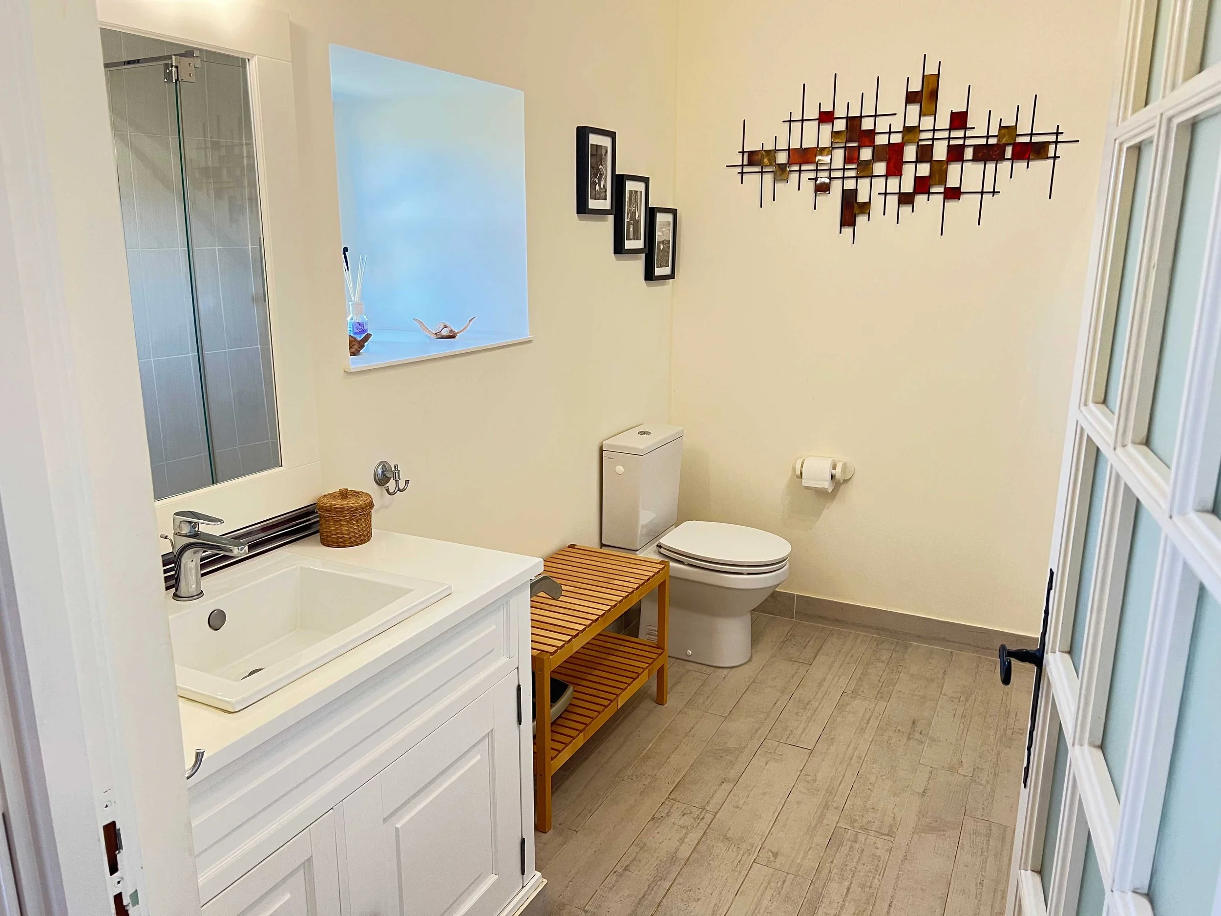 A bathroom with a white vanity sink, a mirror, framed photographs on the wall, a small wicker basket, a wooden bench, a toilet, and a walk-in shower with glass door. The bathroom has beige walls and tile flooring.
