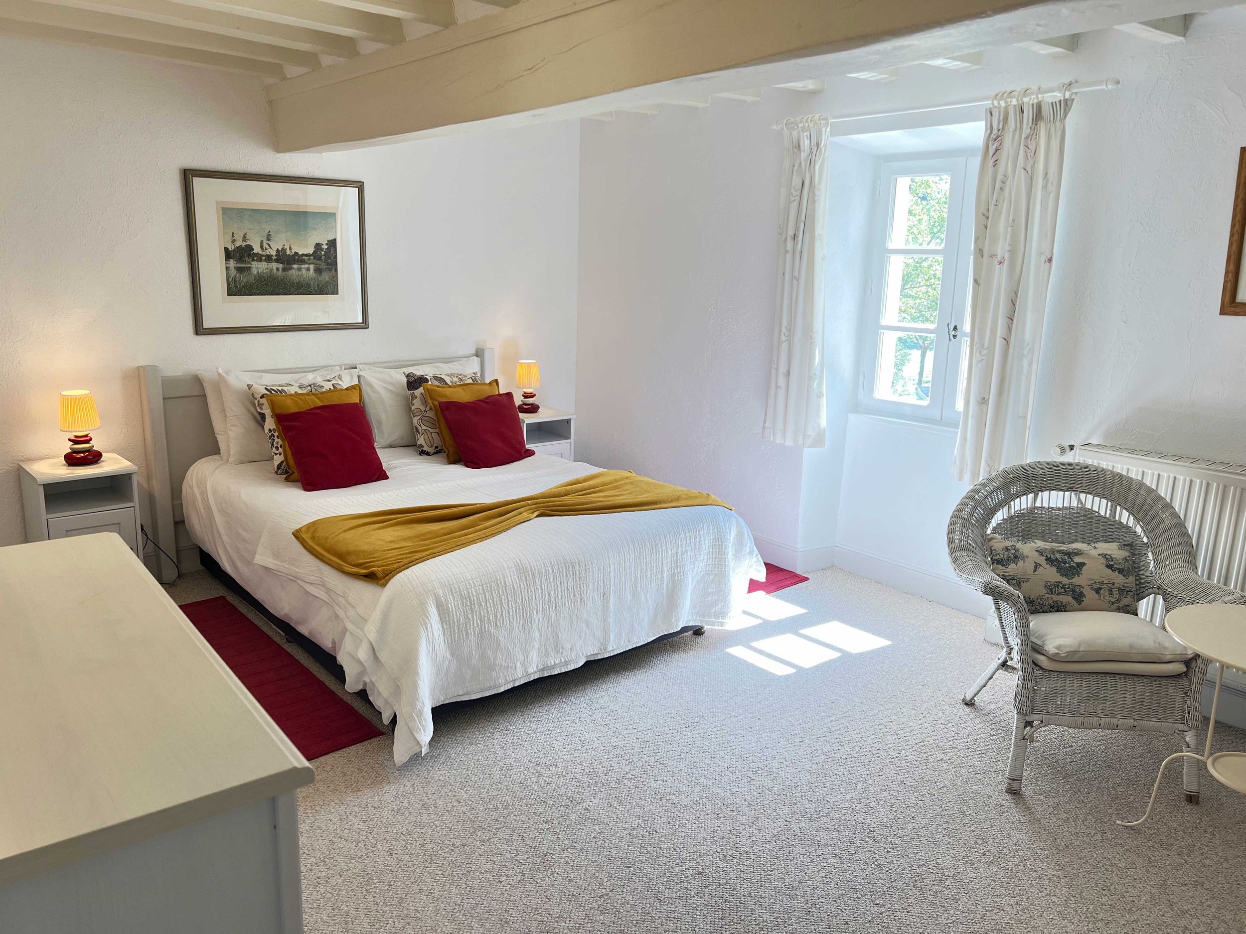 A cozy bedroom with a white bed, red and mustard pillows, and a mustard throw blanket. Two bedside lamps with yellow shades are on white nightstands. A framed picture hangs above the bed. There is a window with white curtains, and a wicker chair with a floral cushion sits near the window. The room has white textured walls and a beige carpet.