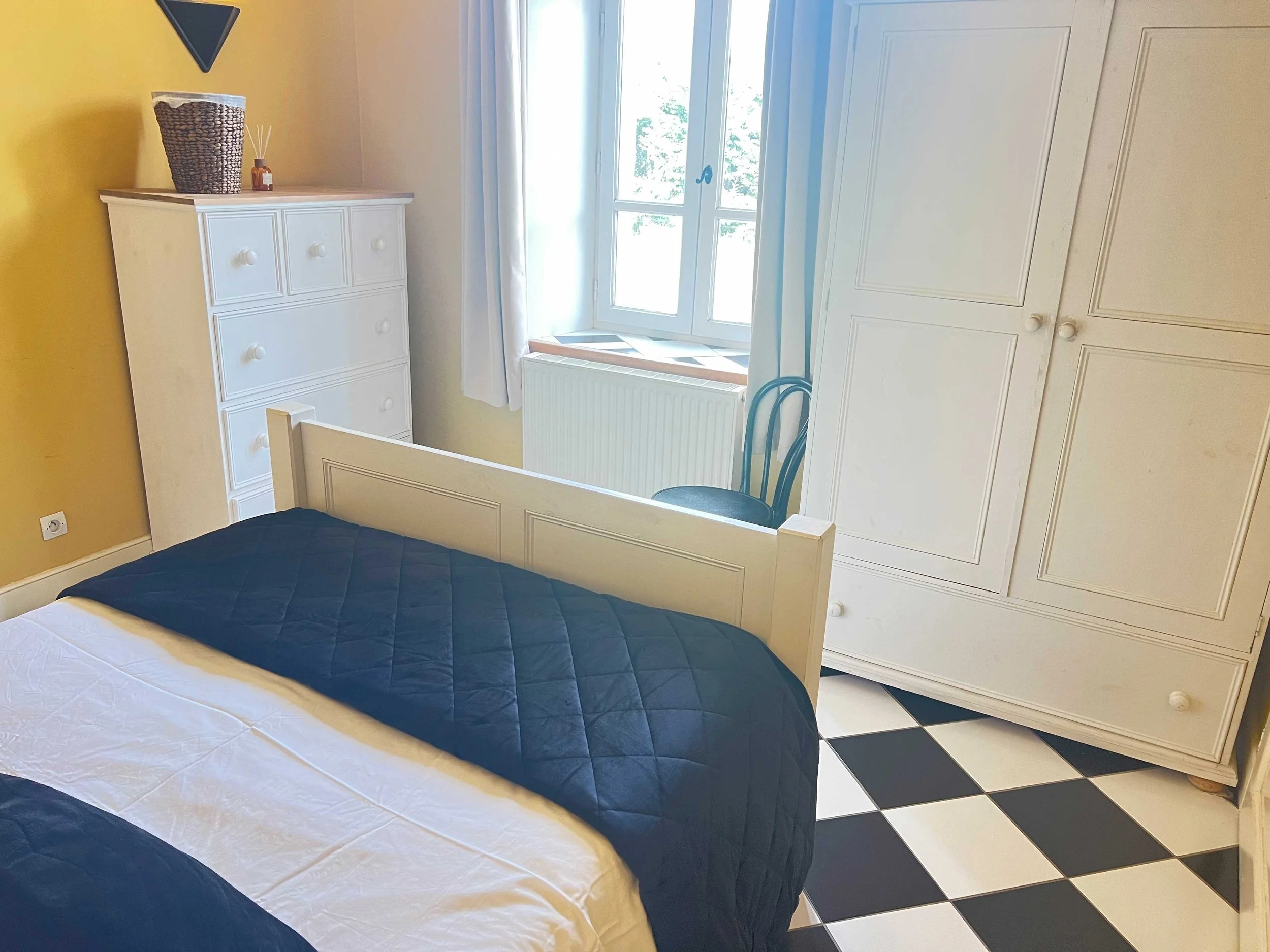 A bedroom with a white bed frame and mattress, a black quilted blanket, a white quilt, a white dresser with drawers, a wicker basket, a small bottle with reed diffusers, a black wall hanging, a large window with white curtains, a white radiator, a black chair, and checkered black and white flooring.