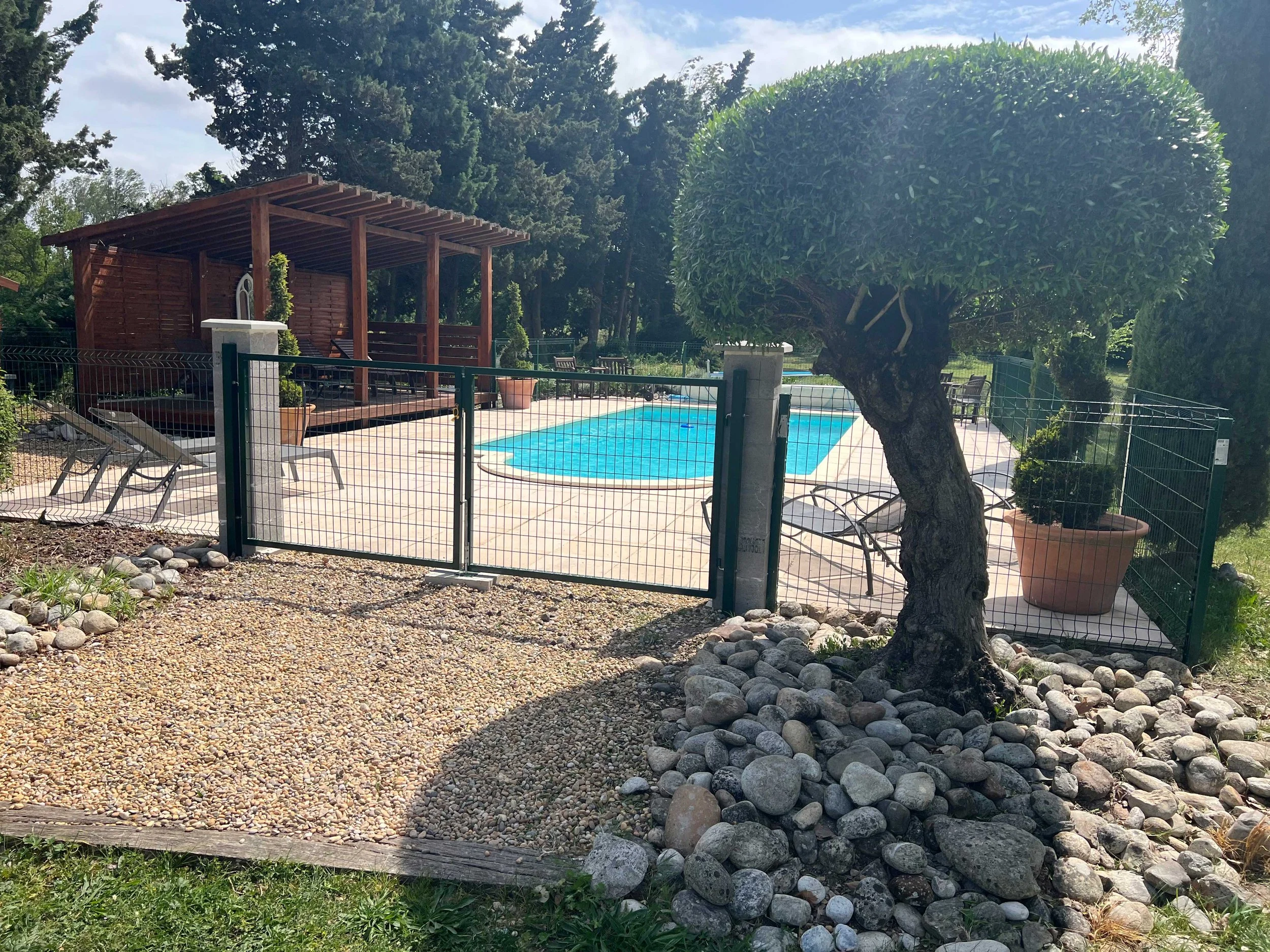 A backyard with a small swimming pool enclosed by a black metal fence. There is a shaded wooden structure with a white fountain and a bench underneath. A large, neatly trimmed, round green tree is in the foreground. Decorated potted plants and outdoor chairs are visible around the pool area. The background includes tall trees and a partly cloudy sky.