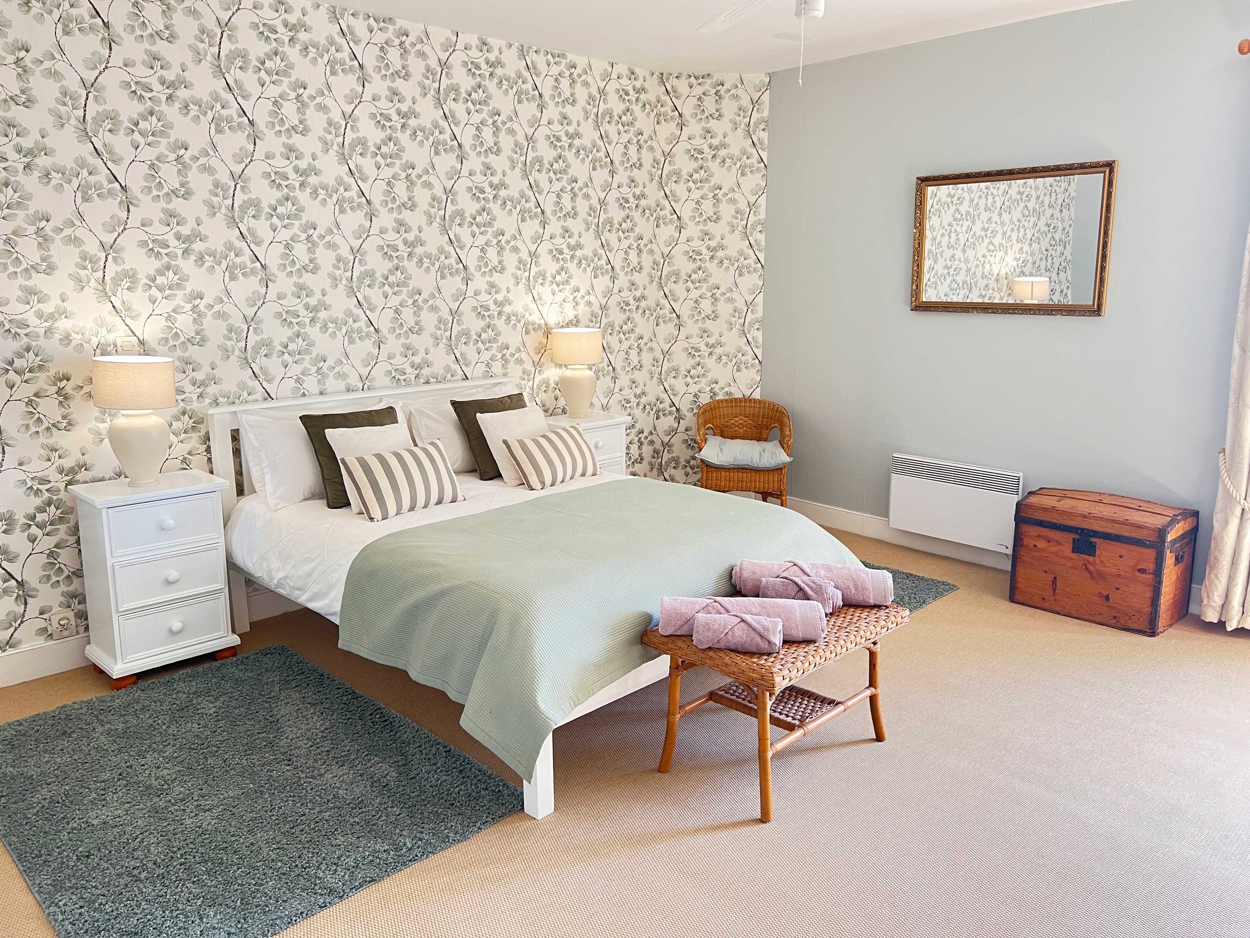 A bedroom with a white bed, striped pillows, a wicker chair, a chest, two white nightstands with lamps, a large mirror with a gold frame, floral wallpaper, and a heater near the corner.